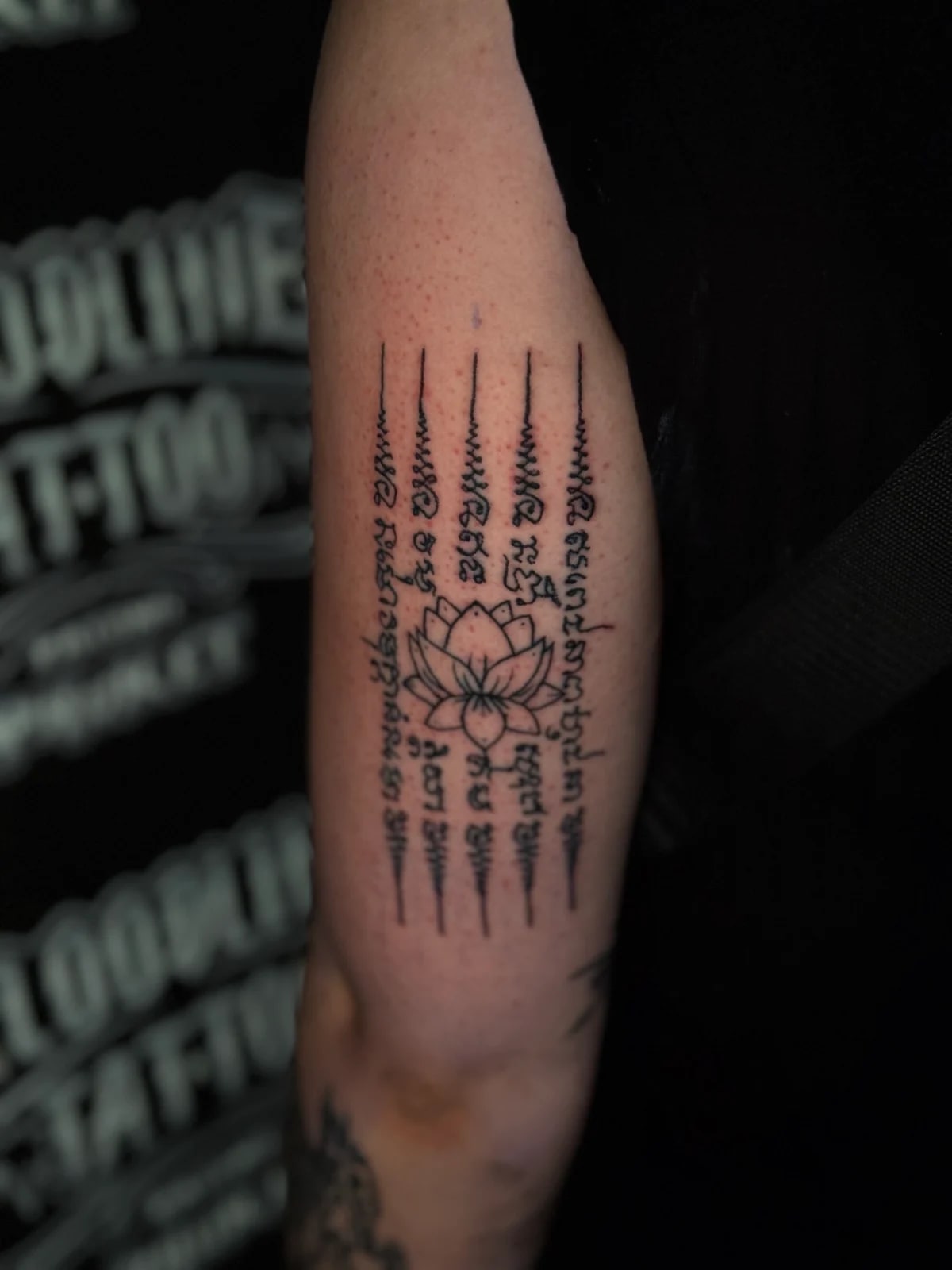 Hah Taew (Five Lines) Sak Yant with five vertical Khom script columns and unalome flourishes framing a central blooming lotus on the upper arm at Bloodline Tattoo Phuket.