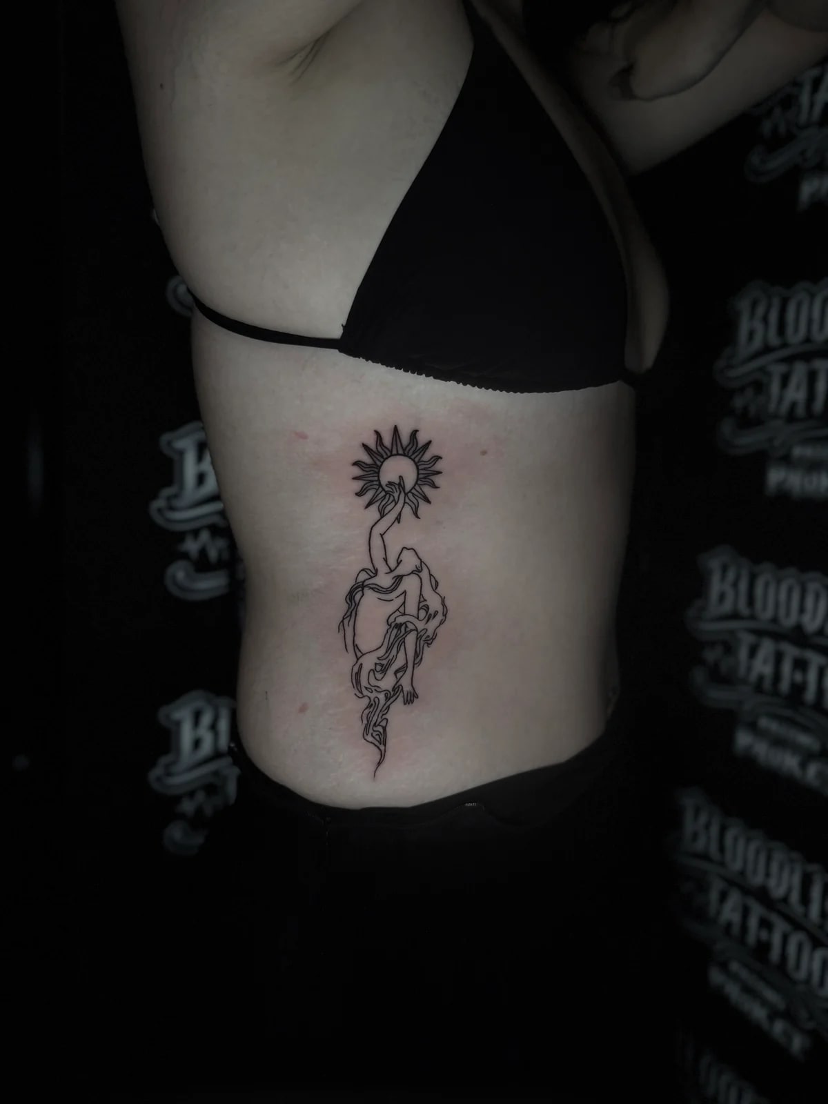 Fine-line bamboo tattoo of a stylized woman reaching toward a radiant sun on the side ribs at Bloodline Tattoo Phuket.