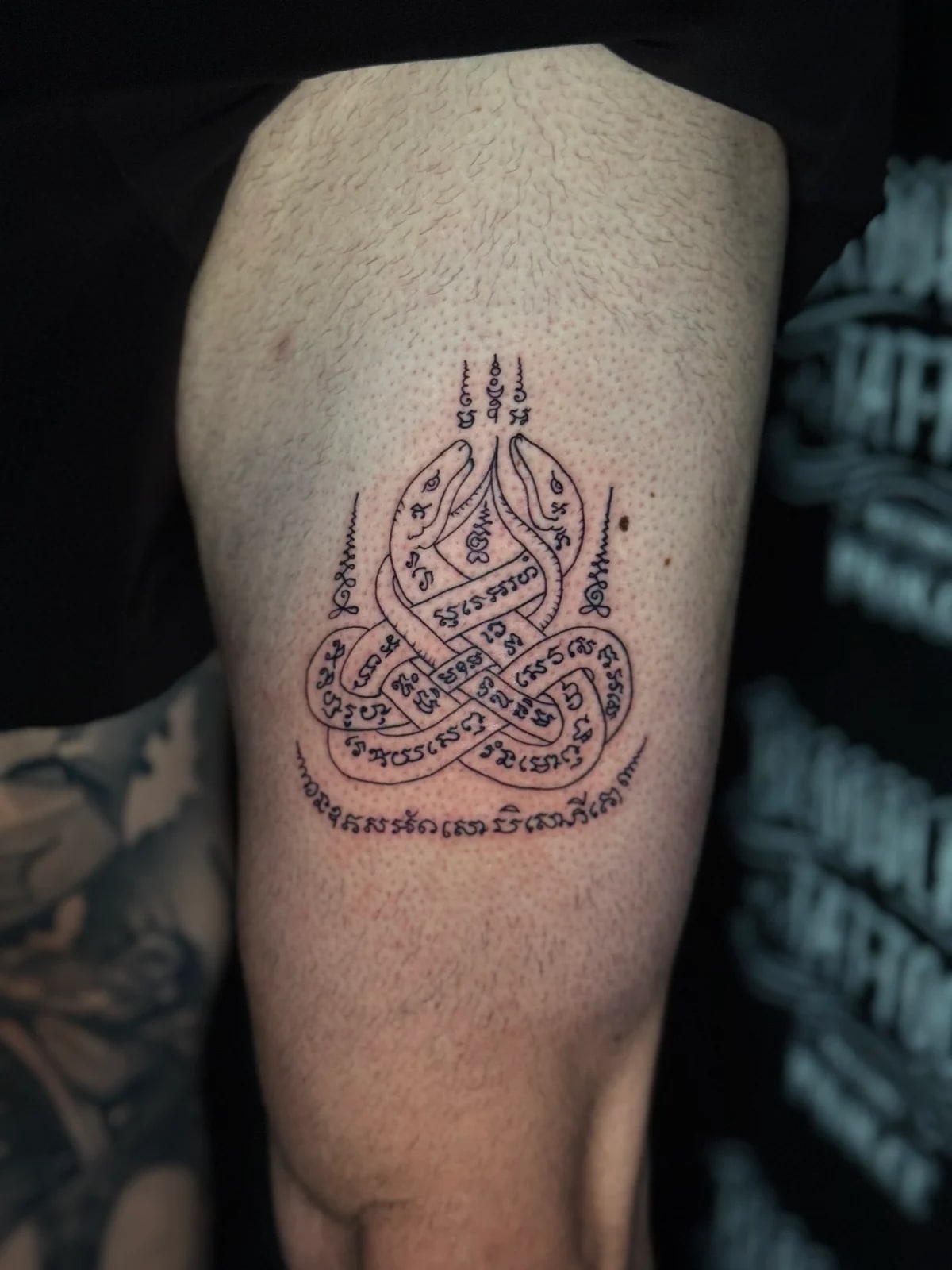 Twin Naga snakes Sak Yant with intertwined serpents, Khom script bodies, and unalome spires on the upper arm at Bloodline Tattoo Phuket.