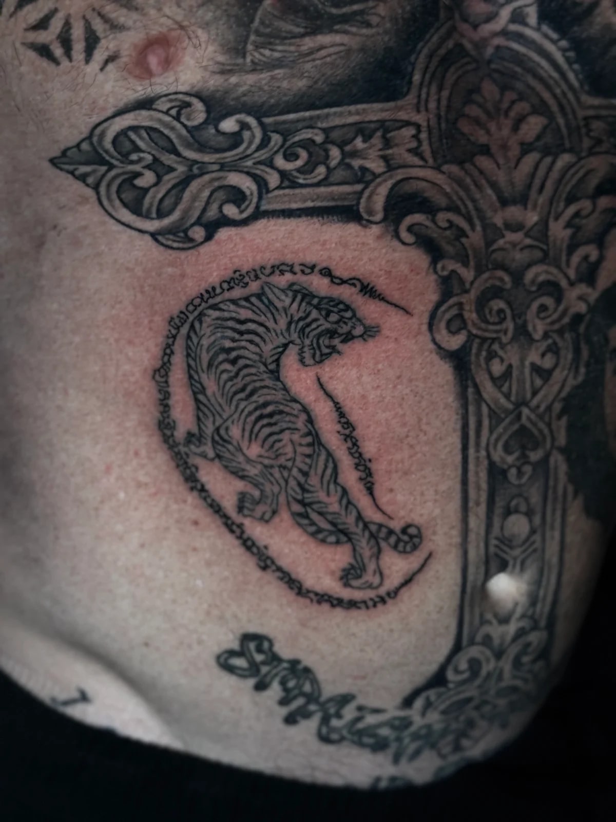 Suea (Tiger) Sak Yant of a snarling tiger with Khom script on the ribs beside an ornate cross tattoo at Bloodline Tattoo Phuket.