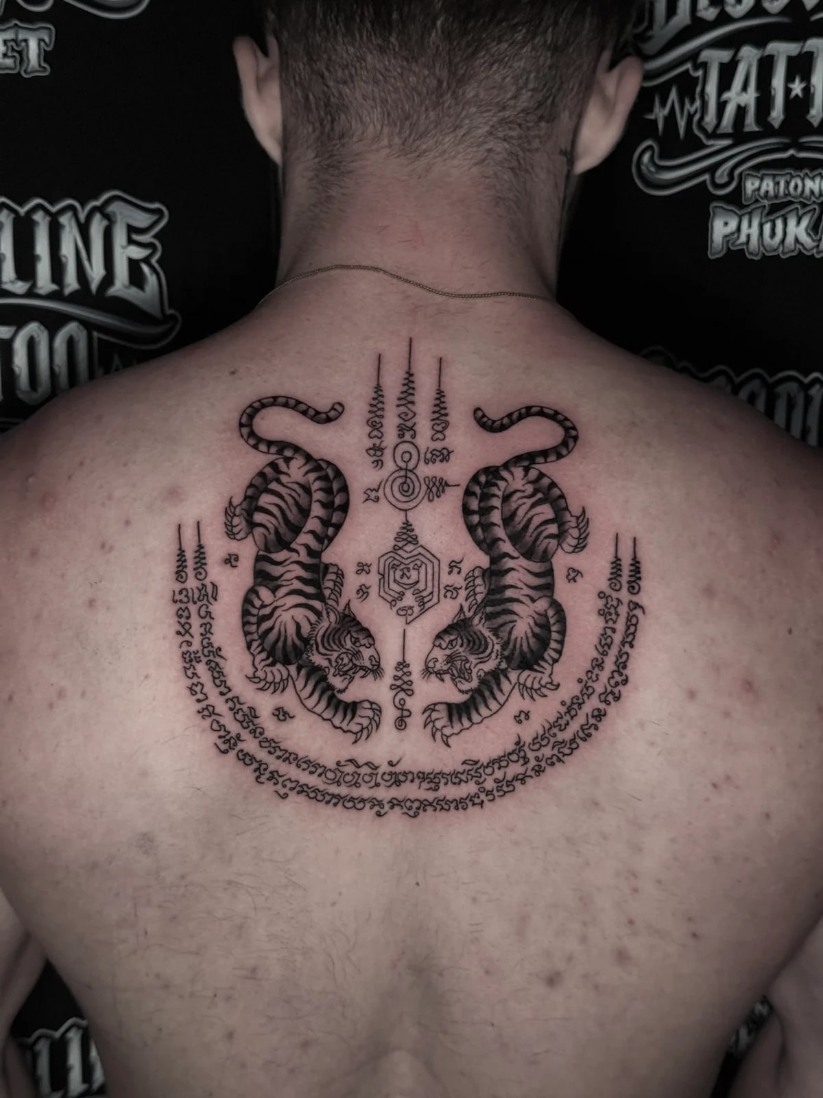 Suea Koo (Twin Tigers) Sak Yant in black and grey with symmetrical tigers, central unalomes, and Khom script crescent on the upper back at Bloodline Tattoo Phuket.