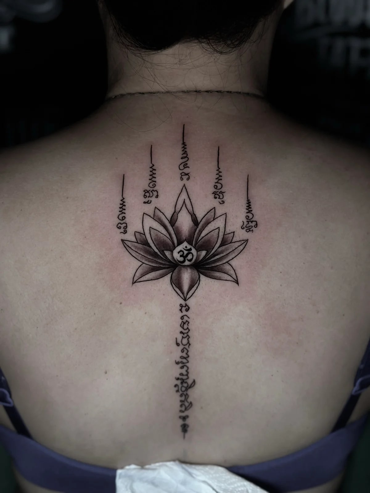 Lotus and Om Sak Yant with shaded lotus petals, five Khom script columns with unalomes, and a spine script line on the upper back at Bloodline Tattoo Phuket.