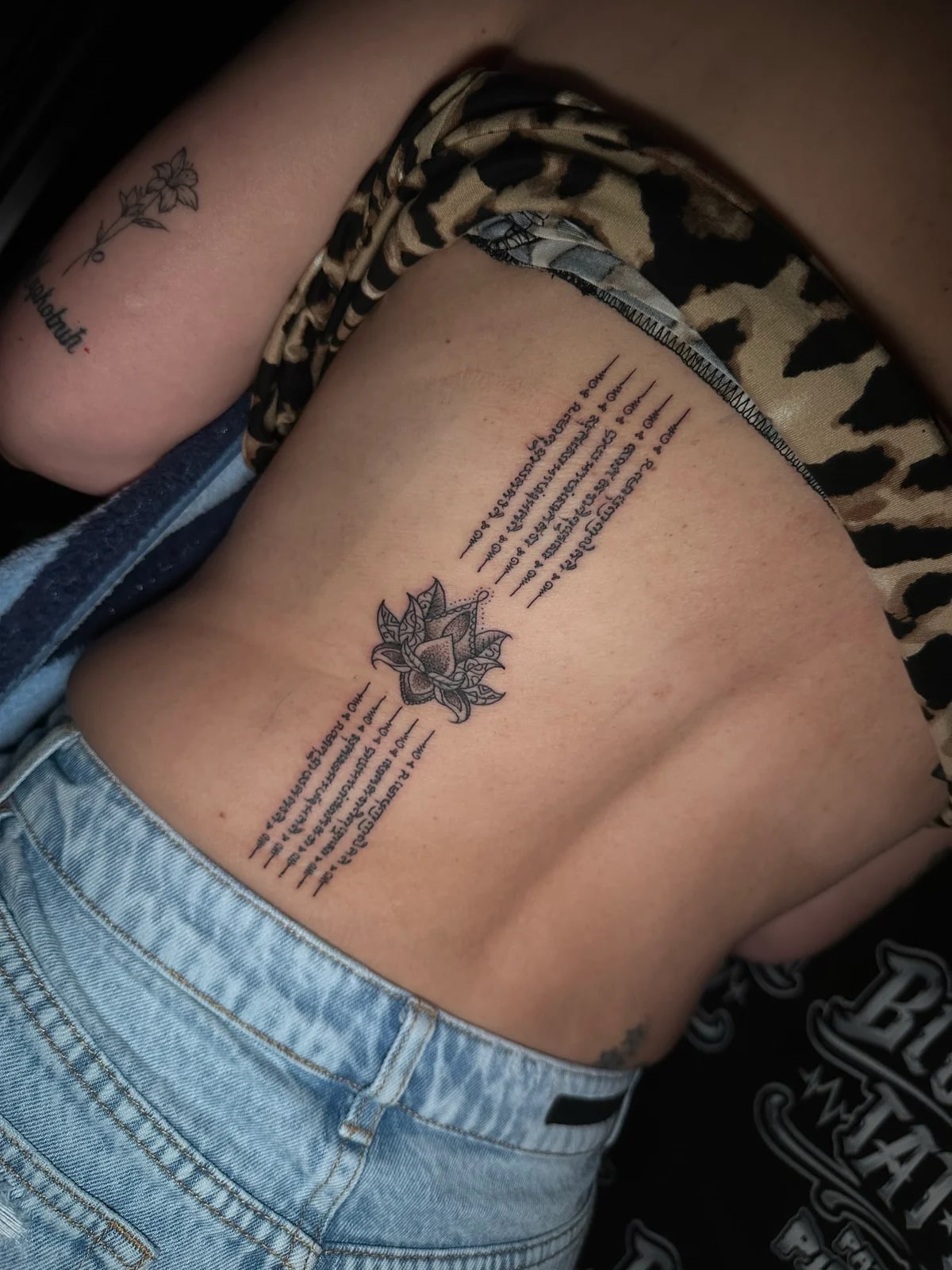Hah Taew-style Sak Yant with a central lotus flower and five vertical Khom script columns along the lower back spine at Bloodline Tattoo Phuket.