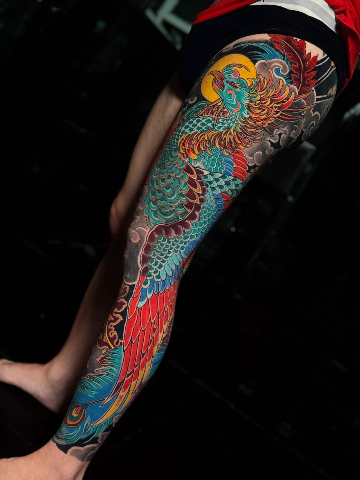 Japanese colour phoenix full-leg tattoo with teal scales, red and yellow tail feathers, and dark cloud backgrounds at Bloodline Tattoo Phuket.