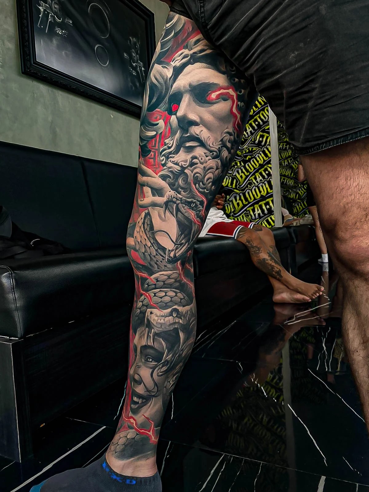Healed black-and-grey leg sleeve with a bearded god, Medusa face, and snake scales, lifted by red lightning accents and crisp healed detail at Bloodline Tattoo Bali.