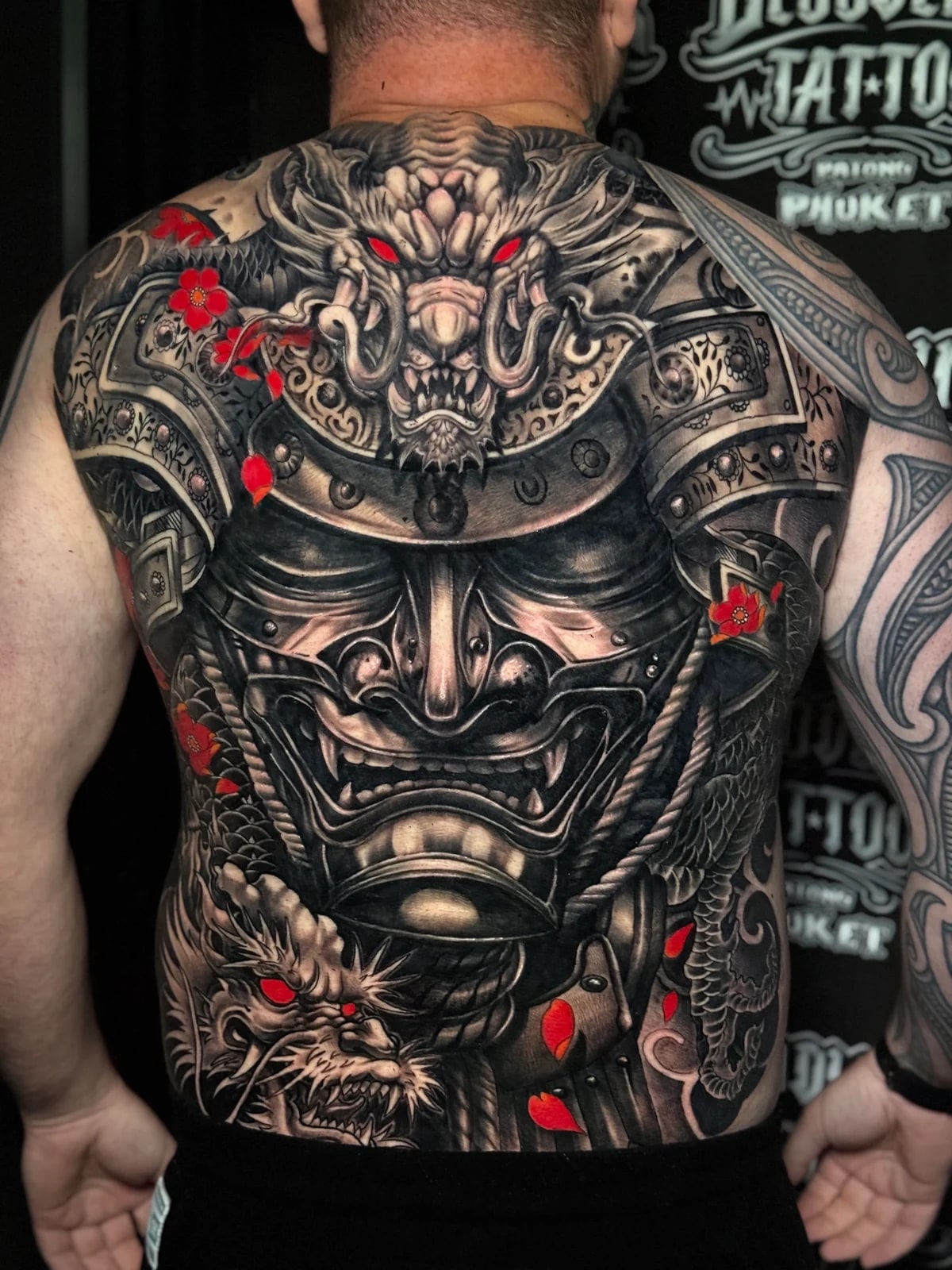 Japanese black and grey full-back tattoo with samurai menpo and kabuto, flanking dragon heads with red eyes, and red cherry blossoms at Bloodline Tattoo Phuket.