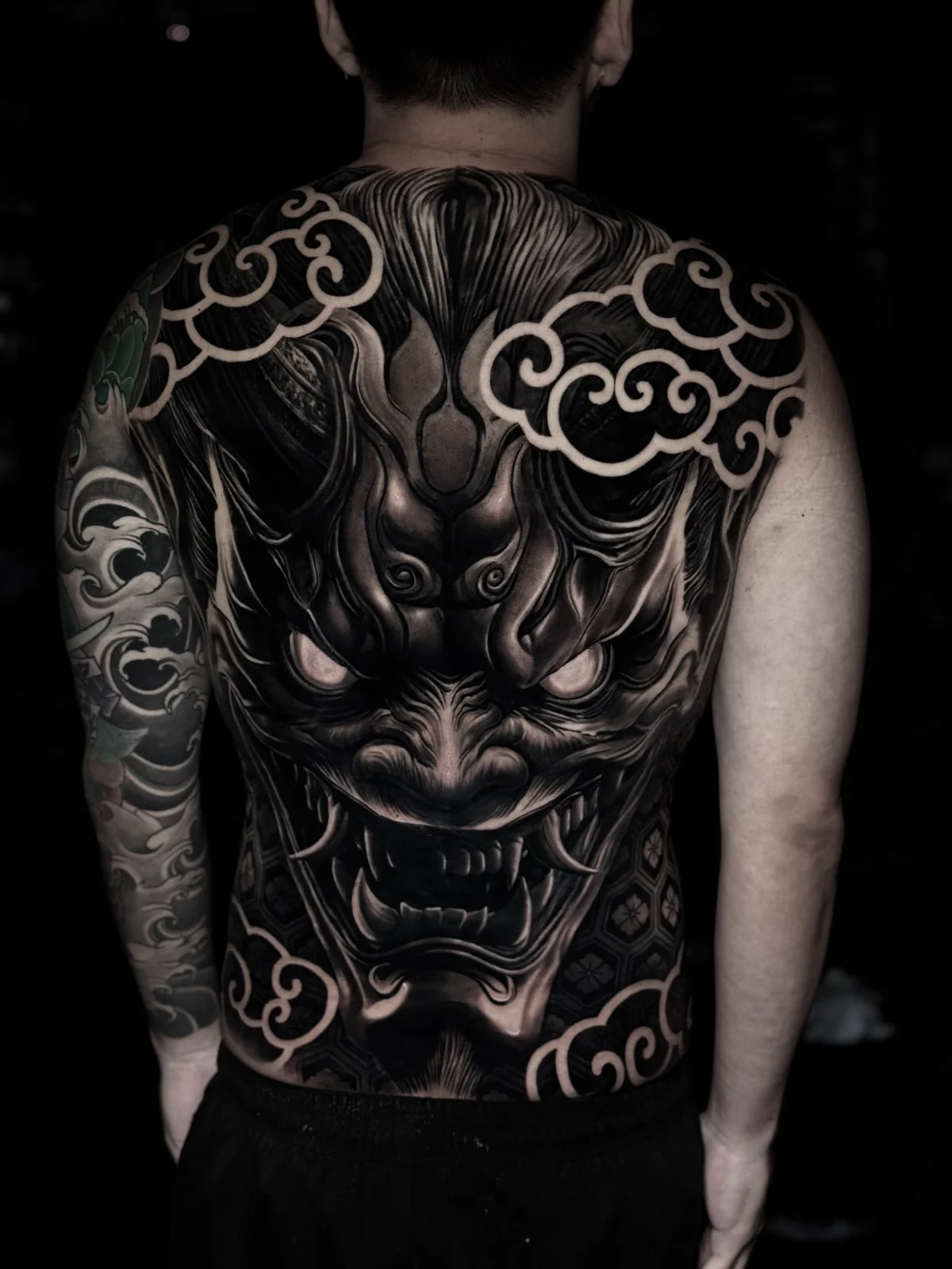 Japanese black and grey full-back Hannya mask with glowing eyes, sharp fangs, and stylized cloud wind bars at Bloodline Tattoo Phuket.