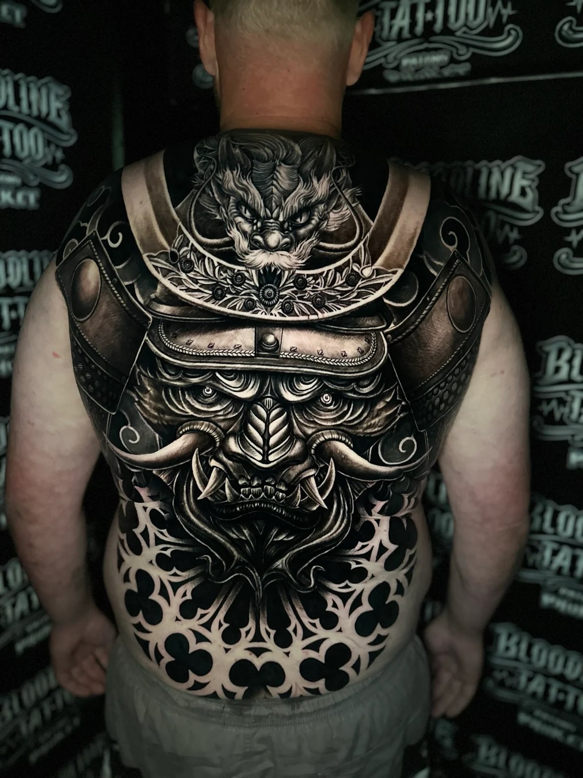 Japanese black and grey full-back tattoo with shishi head at the neck, ornate samurai menpo and kabuto, and bold geometric fill on the lower back at Bloodline Tattoo Phuket.