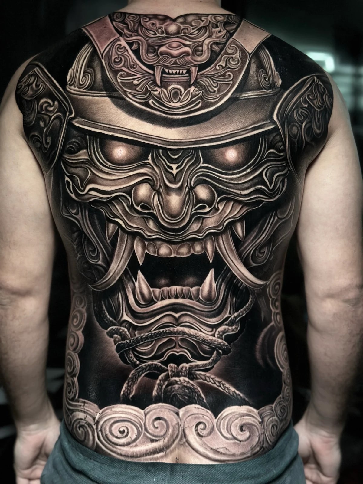 Japanese black and grey full-back samurai menpo with kabuto crest, braided ropes, and stylized clouds on a solid black ground at Bloodline Tattoo Phuket.