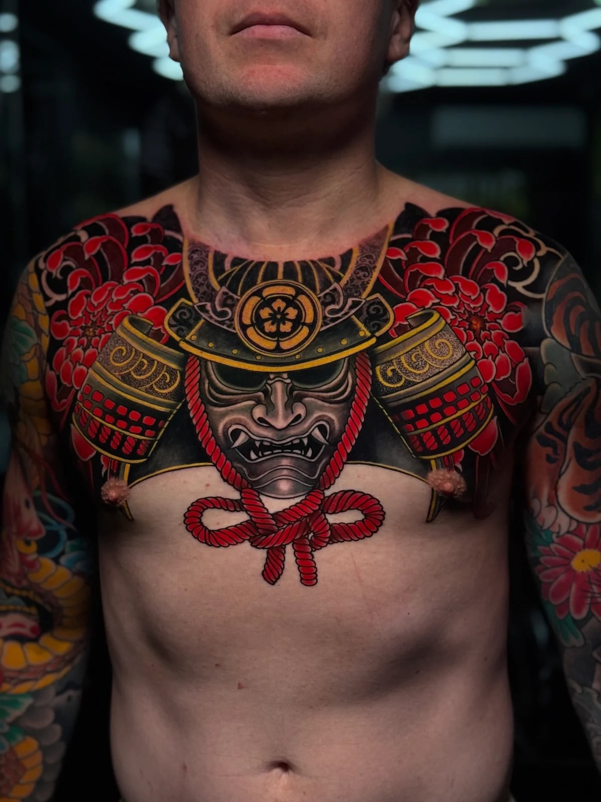 Japanese chest piece with samurai menpo, gold-accent kabuto, red agemaki knot, and vivid red chrysanthemums at Bloodline Tattoo Phuket.