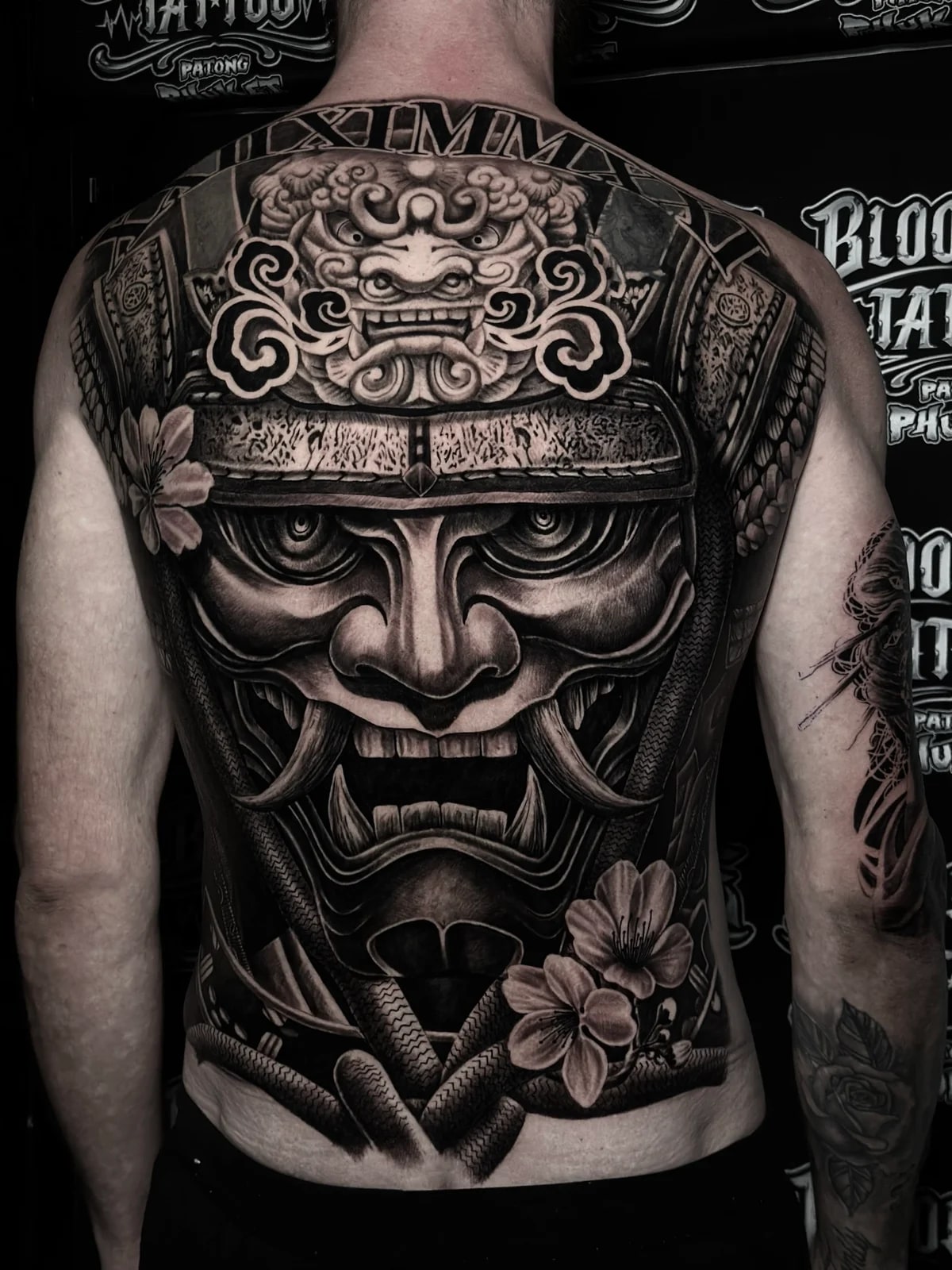 Japanese black and grey full-back samurai mask with shishi crest, cherry blossoms, and roman numerals at the neckline at Bloodline Tattoo Phuket.