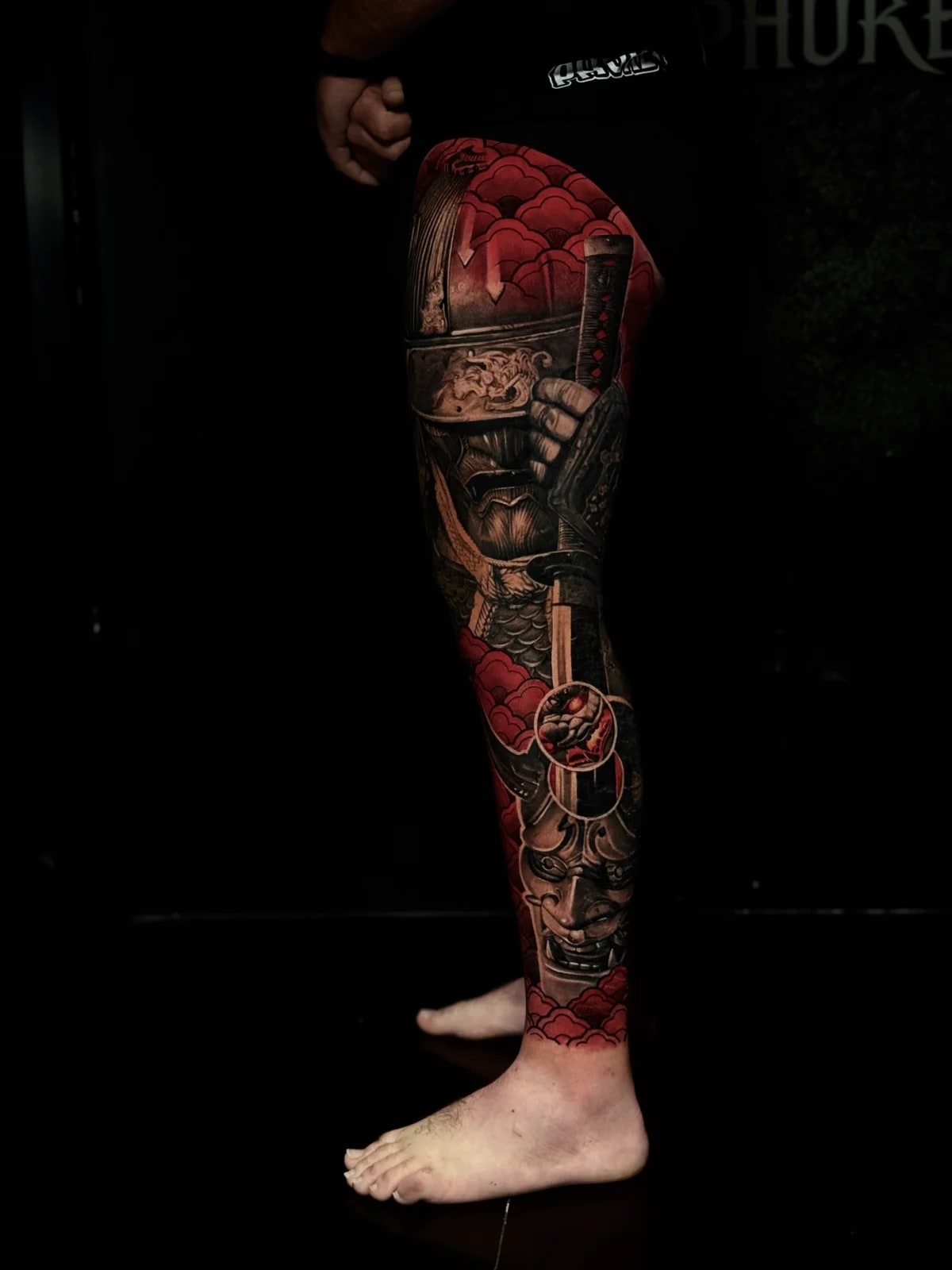 Japanese full leg sleeve with armored samurai holding a katana on the thigh and Hannya mask on the calf amid vibrant red clouds at Bloodline Tattoo Phuket.