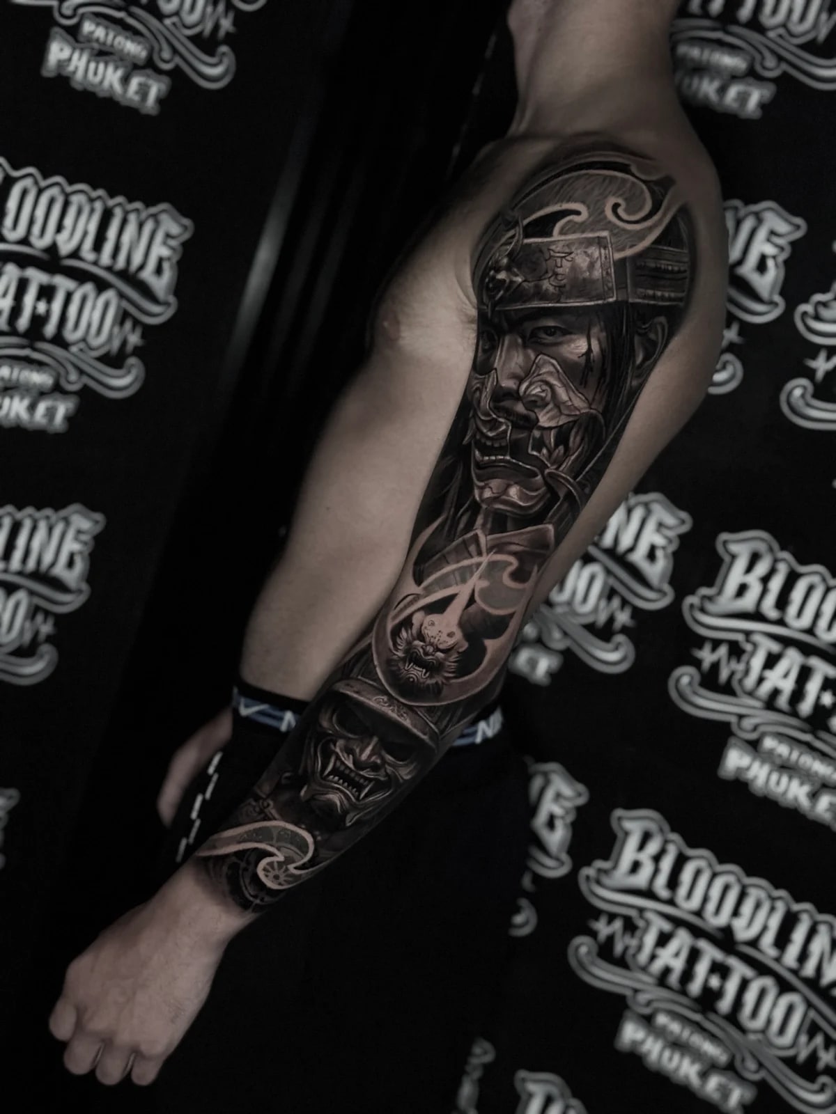 Japanese black and grey full sleeve with samurai in kabuto and menpo above a fierce Hannya mask amid stylized waves at Bloodline Tattoo Phuket.