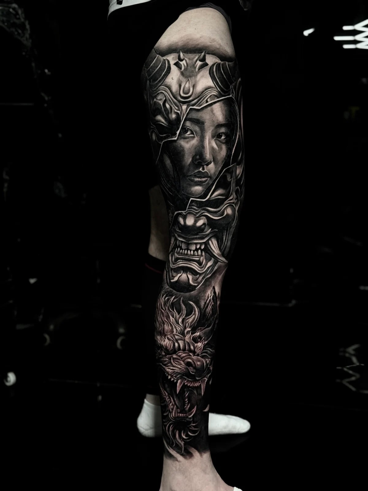 Japanese black and grey full leg tattoo with woman's face through a cracked Hannya mask on the thigh and dragon head on the shin at Bloodline Tattoo Phuket.
