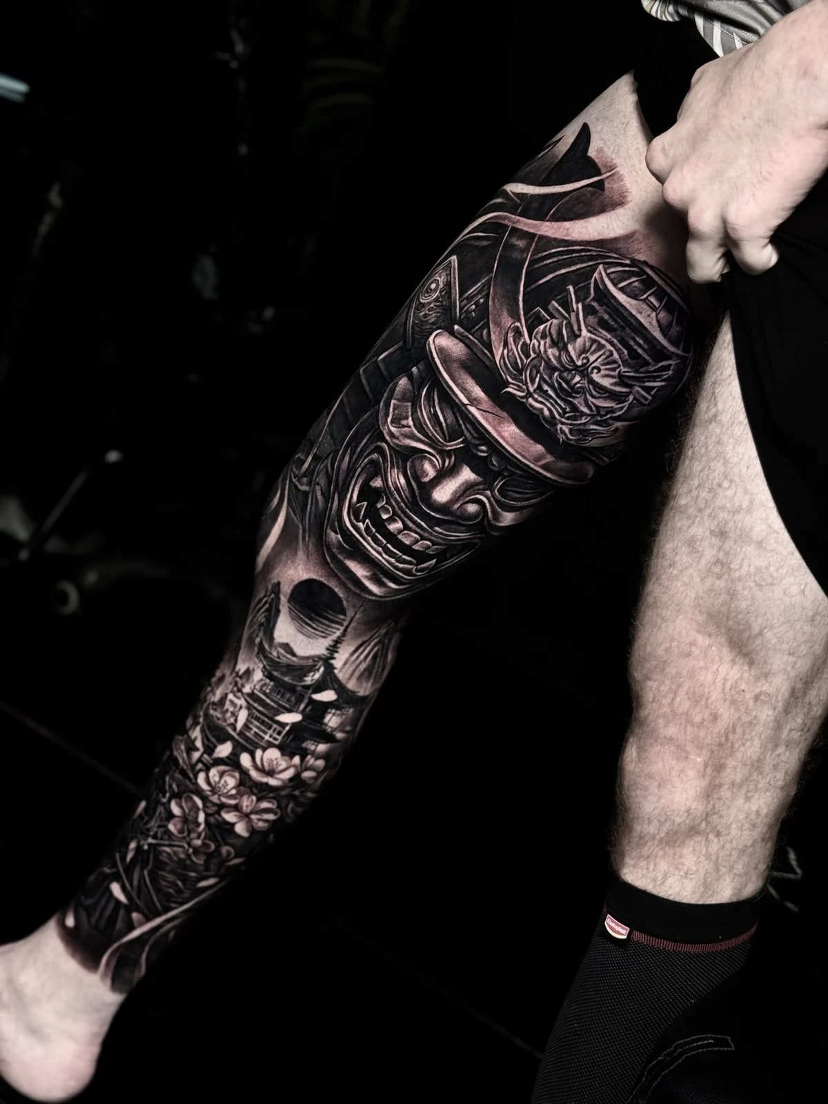 Japanese black and grey leg sleeve with fierce samurai menpo and kabuto, pagoda, cherry blossoms, and dark sun disc at Bloodline Tattoo Phuket.