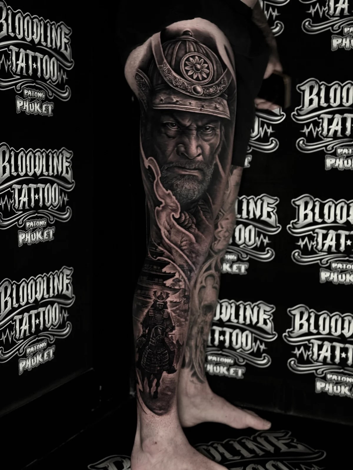 Japanese black and grey full leg tattoo with stern samurai portrait in kabuto on the thigh and armored samurai on horseback with pagoda below at Bloodline Tattoo Phuket.