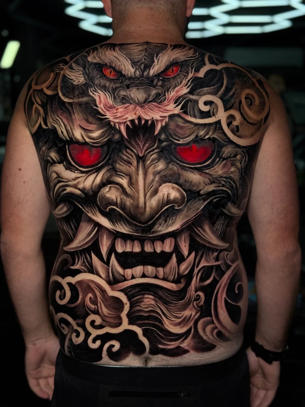 Japanese full-back Hannya mask with glowing red eyes, horned creature crest, and stylized clouds in black and grey at Bloodline Tattoo Phuket.