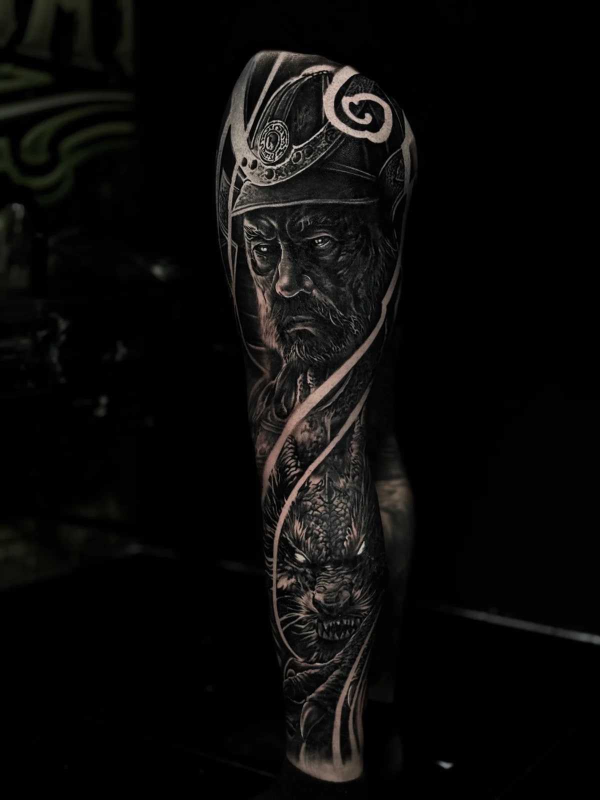 Japanese black and grey full leg tattoo with bearded elder in kabuto on the thigh and dragon head with glowing eyes amid bold white wind bars at Bloodline Tattoo Phuket.