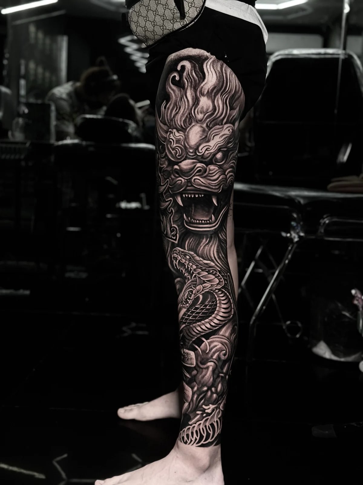Japanese black and grey full leg sleeve with snarling foo dog on the thigh and coiled snake on the calf amid clouds and waves at Bloodline Tattoo Phuket.