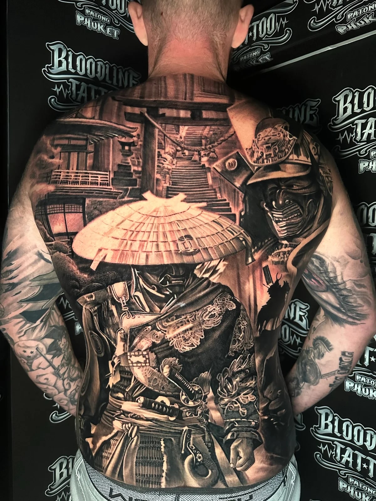 Japanese black and grey full-back with ronin in straw hat and mempo, torii gate and temple stairs, and large kabuto with mounted samurai at Bloodline Tattoo Phuket.