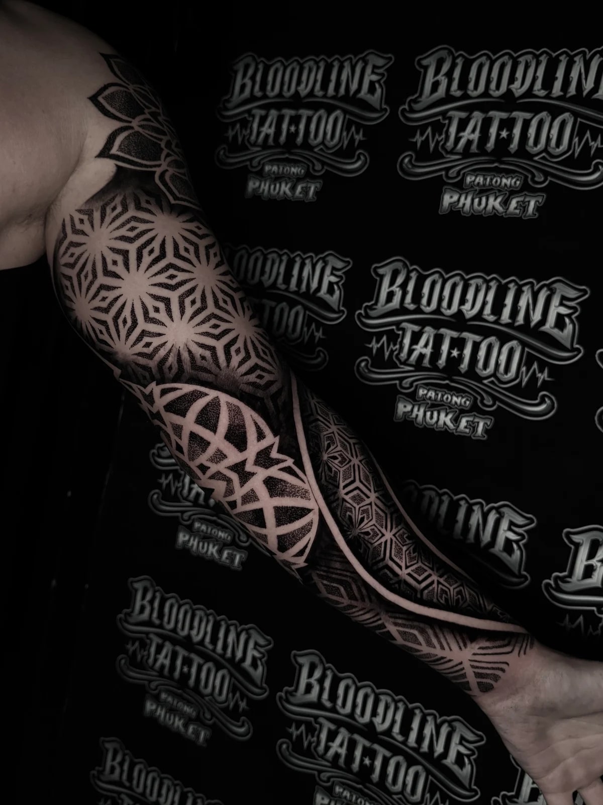 Geometric mandala sleeve with star-shaped upper arm mandalas, a sweeping negative-space curve, and stippled hexagonal wrist fill at Bloodline Tattoo Phuket.