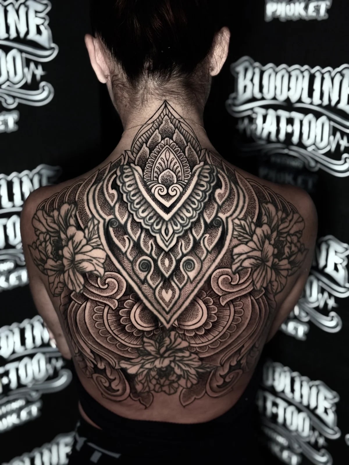 Symmetrical ornamental back mandala with a diamond central motif, stippled dotwork shading, and large peony florals on the shoulder blades at Bloodline Tattoo Phuket.
