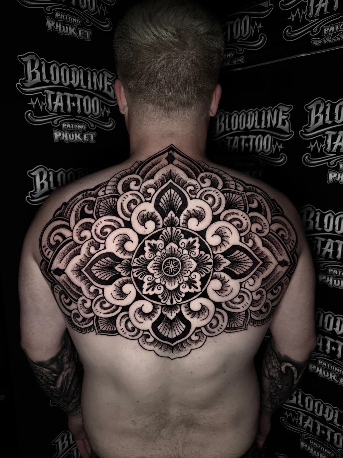 Upper-back symmetrical mandala with ornate swirling layers, bold black outlines, and fine stippled dotwork shading at Bloodline Tattoo Phuket.