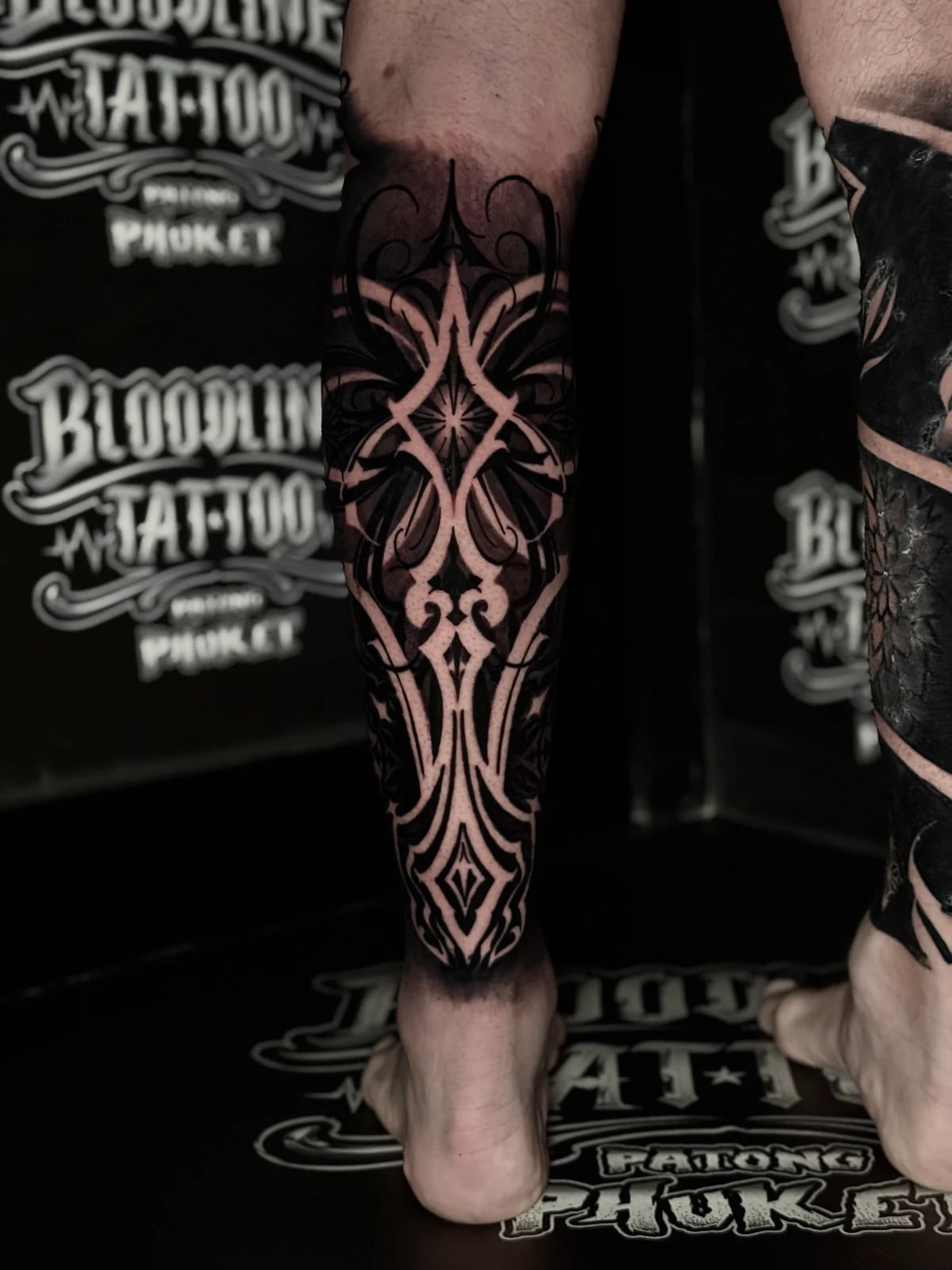 Ornamental blackwork calf tattoo with a symmetrical diamond sunburst and mirrored blade-like geometric panels at Bloodline Tattoo Phuket.