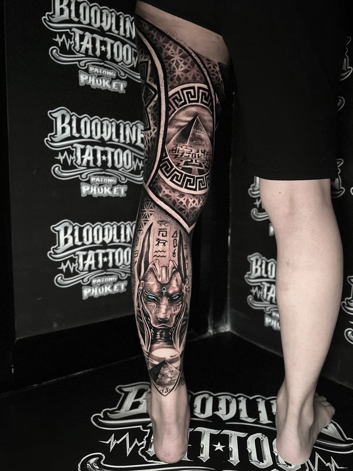 Egyptian-themed leg tattoo with blue-eyed Anubis, hieroglyphs, pyramid emblems, and Flower of Life geometric backgrounds at Bloodline Tattoo Phuket.