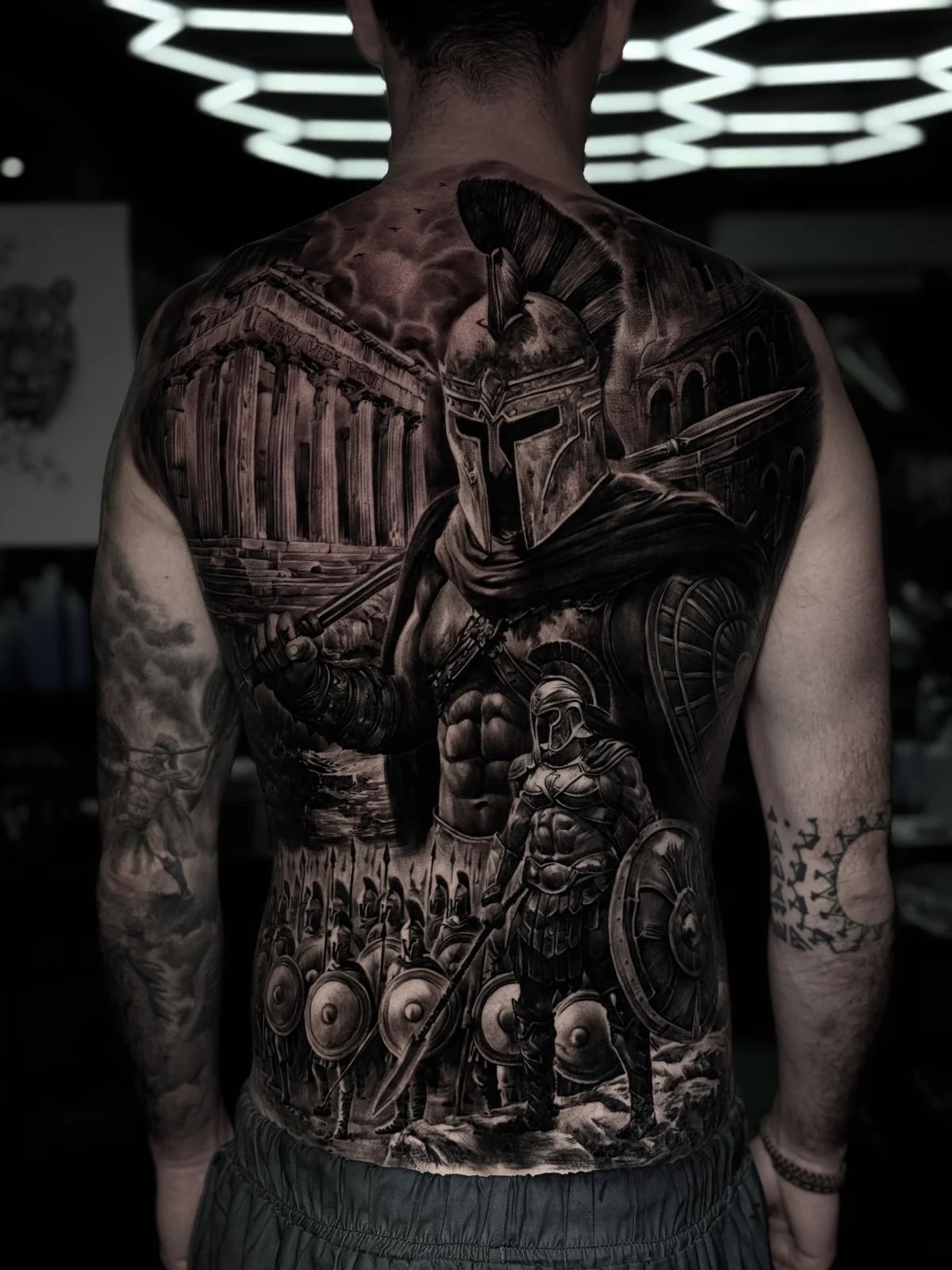 Detailed black and grey realism full back tattoo featuring a Spartan warrior in a Corinthian helmet, Greek temple columns, and a phalanx of shield-bearing soldiers at Bloodline Tattoo Phuket.