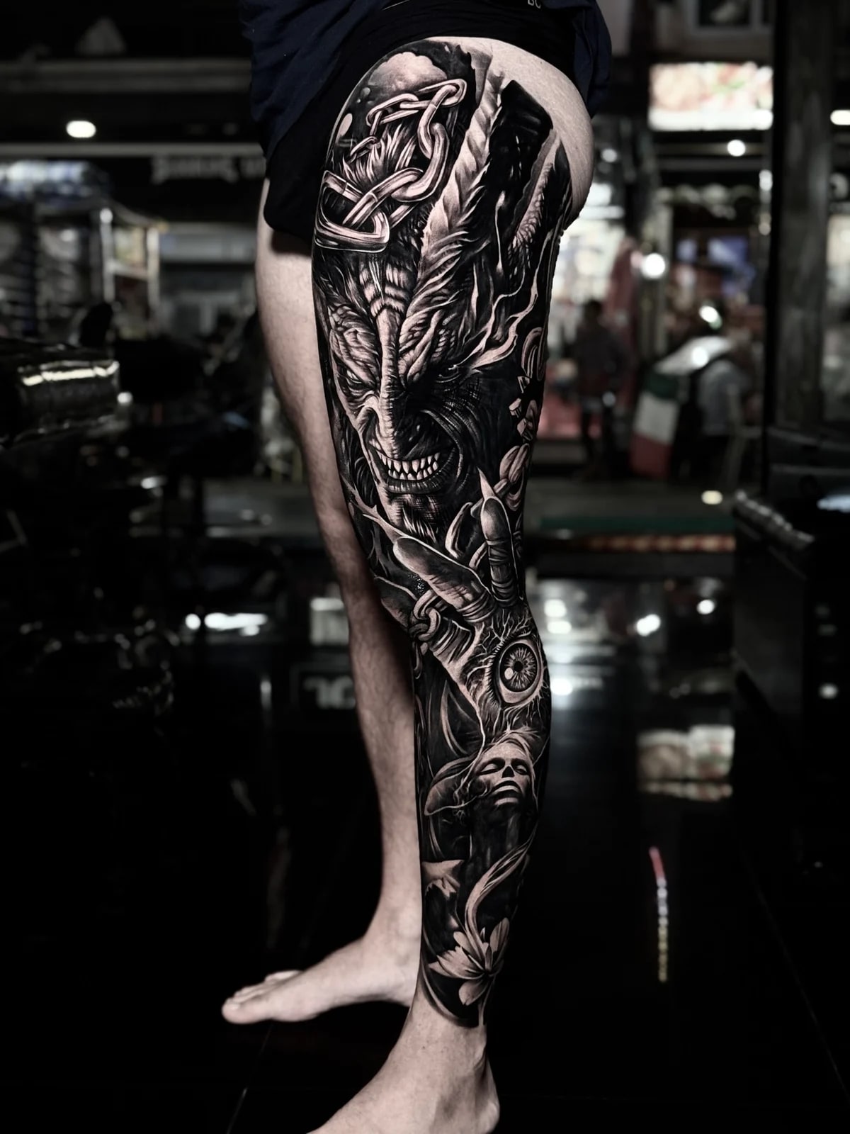 Detailed black and grey realism full leg sleeve tattoo featuring a snarling demon, metal chains, a realistic eye in a clawed hand, a serene woman, and a lotus flower at Bloodline Tattoo Phuket.