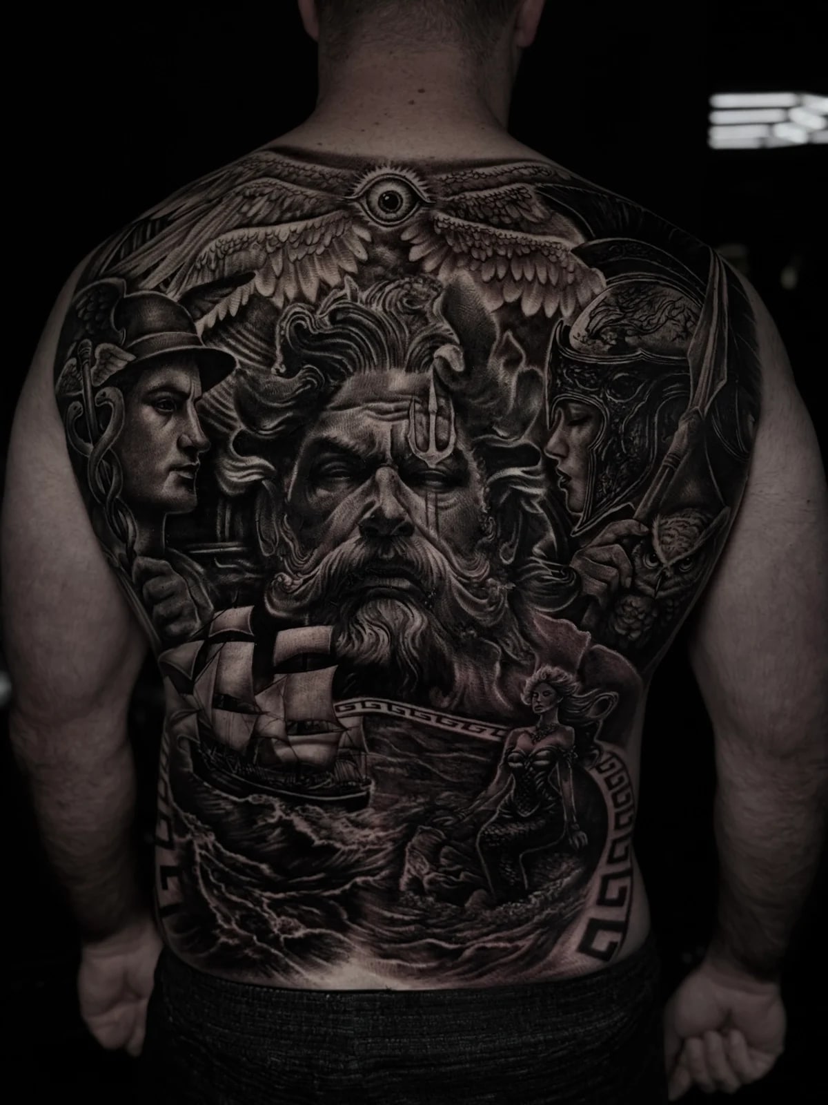 Highly detailed black and grey realism full back tattoo featuring Poseidon, winged all-seeing eye, Hermes, Athena with owl, sailing ship, mermaid, and Greek key border at Bloodline Tattoo Phuket.