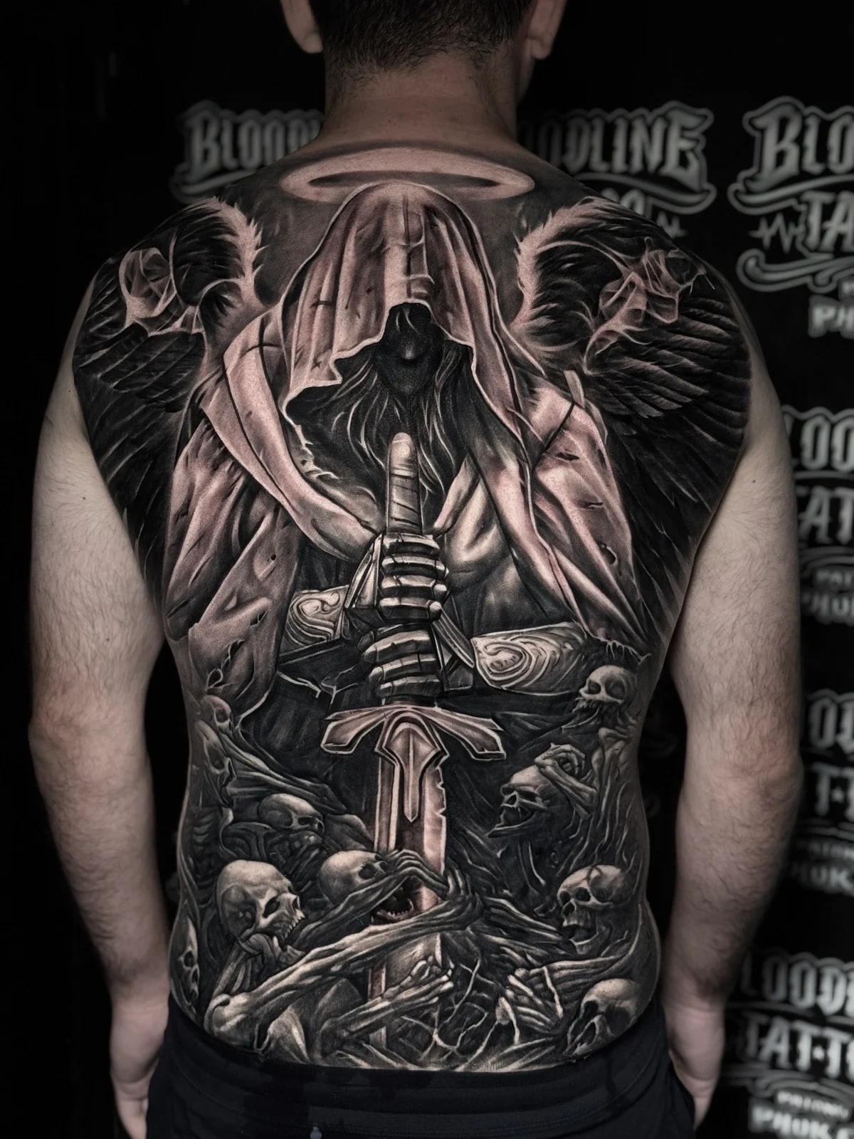 Detailed black and grey realism full back tattoo of a hooded winged angel holding a broadsword above a pile of skulls and reaching skeletal hands at Bloodline Tattoo Phuket.