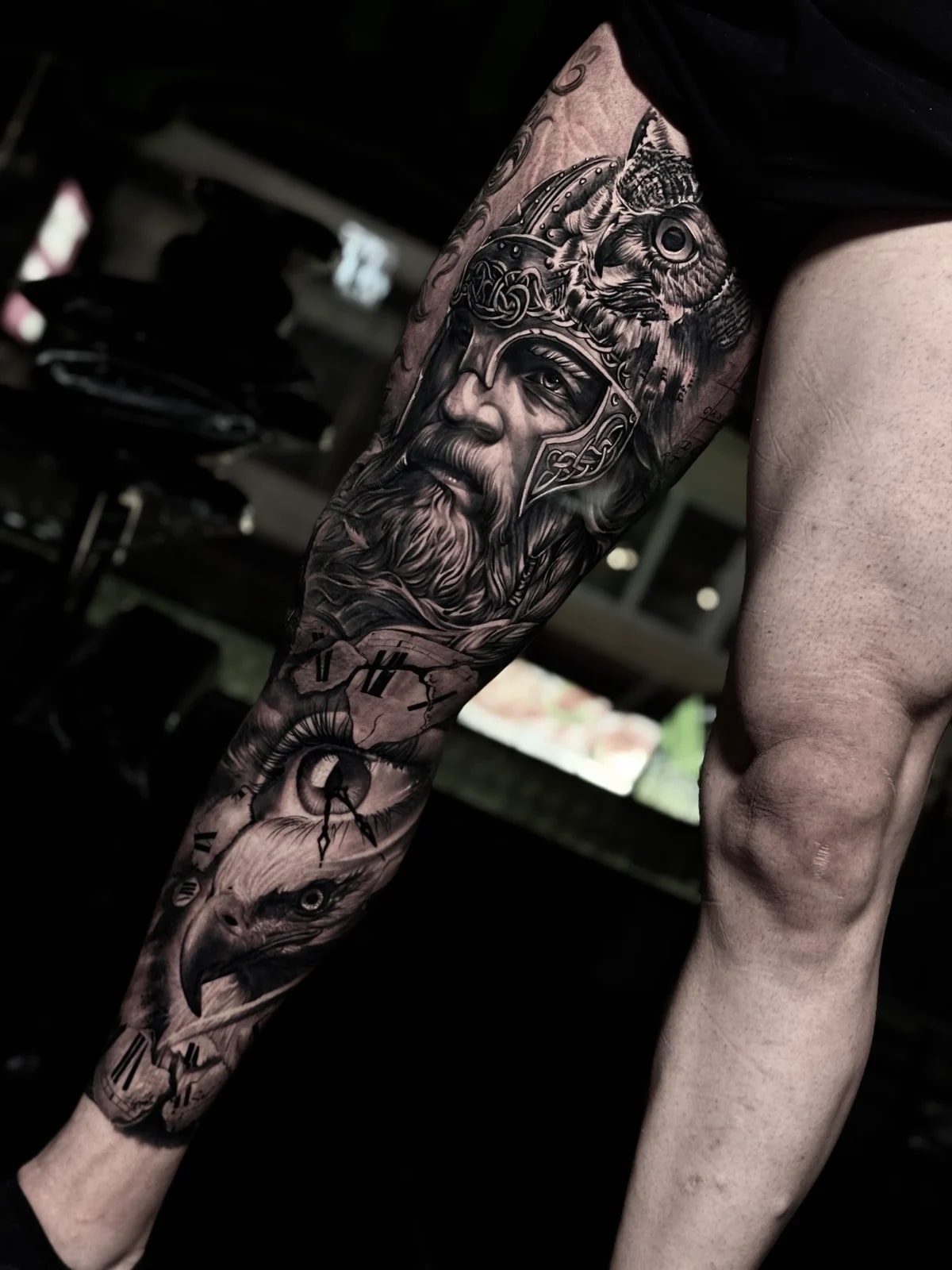 Detailed black and grey realism full leg sleeve tattoo featuring a Viking warrior in a knotwork helmet, an owl, a shattered clock with a realistic eye, and an eagle head at Bloodline Tattoo Phuket.