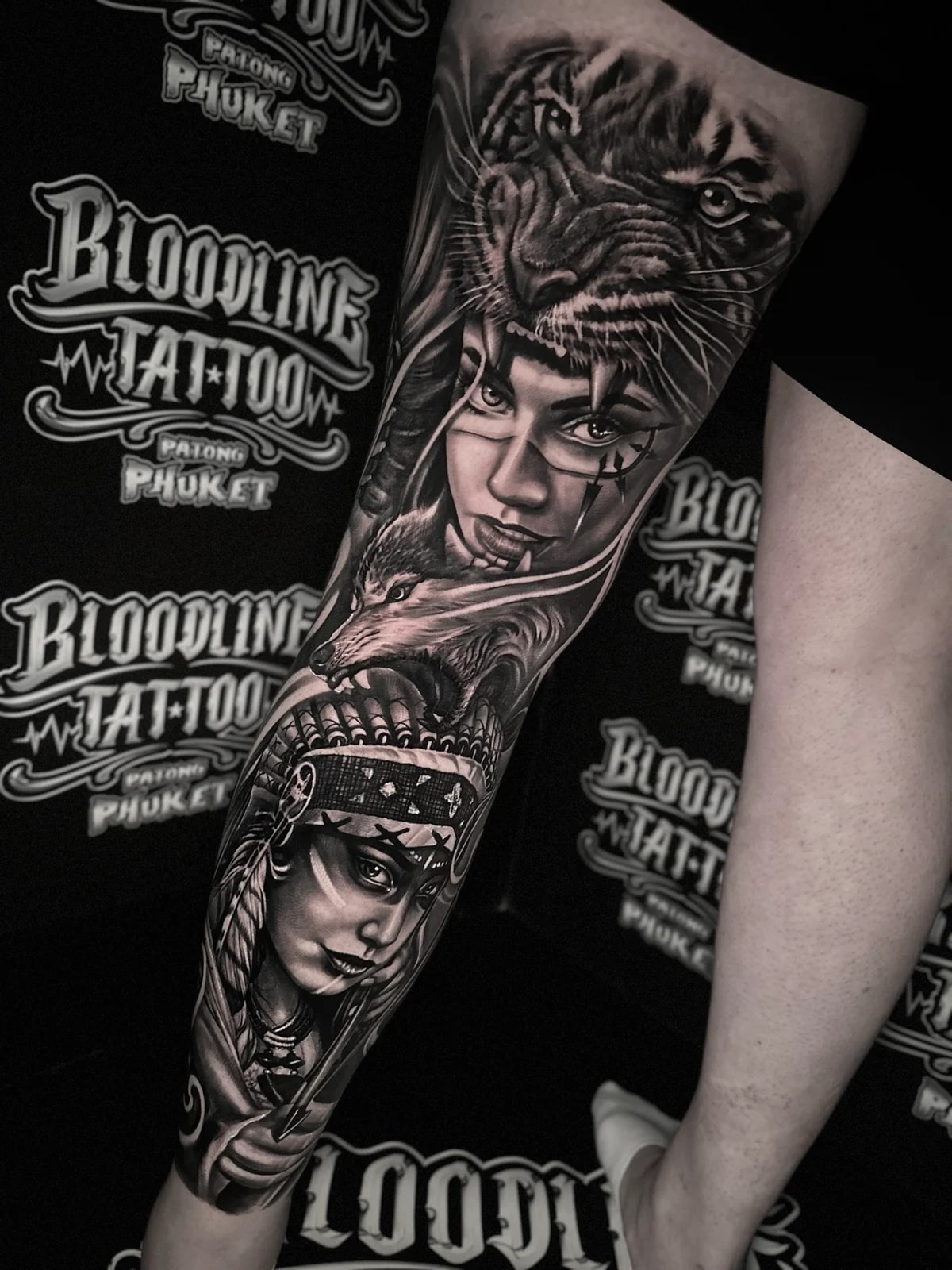 Detailed black and grey realism full leg sleeve tattoo featuring a tiger headdress, tribal-marked woman, wolf profile, and Native American feathered headdress portrait at Bloodline Tattoo Phuket.