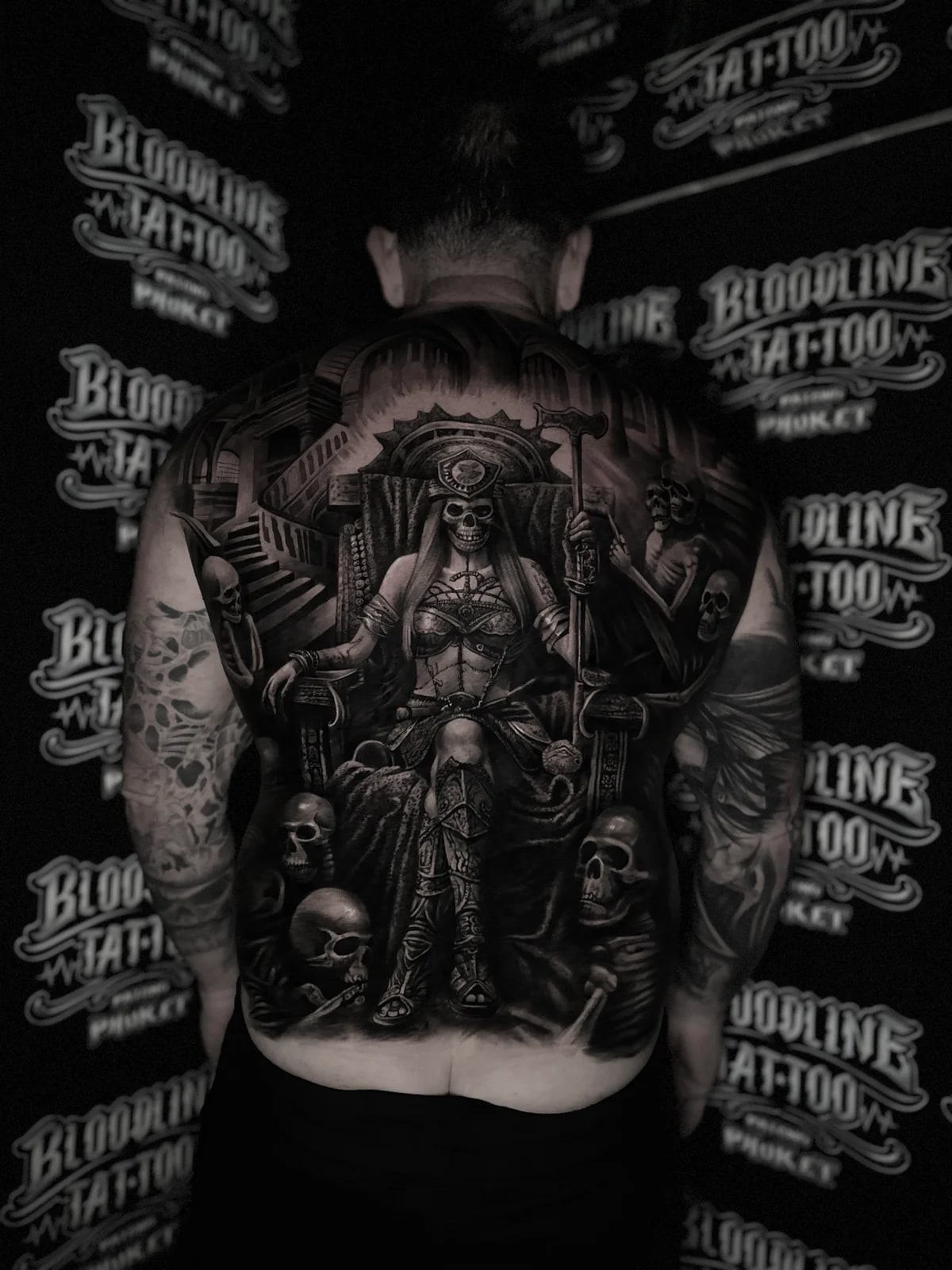 Detailed black and grey realism full back tattoo of a skull-faced warrior woman on a gothic throne with staff, skulls, and skeletal figure at Bloodline Tattoo Phuket.