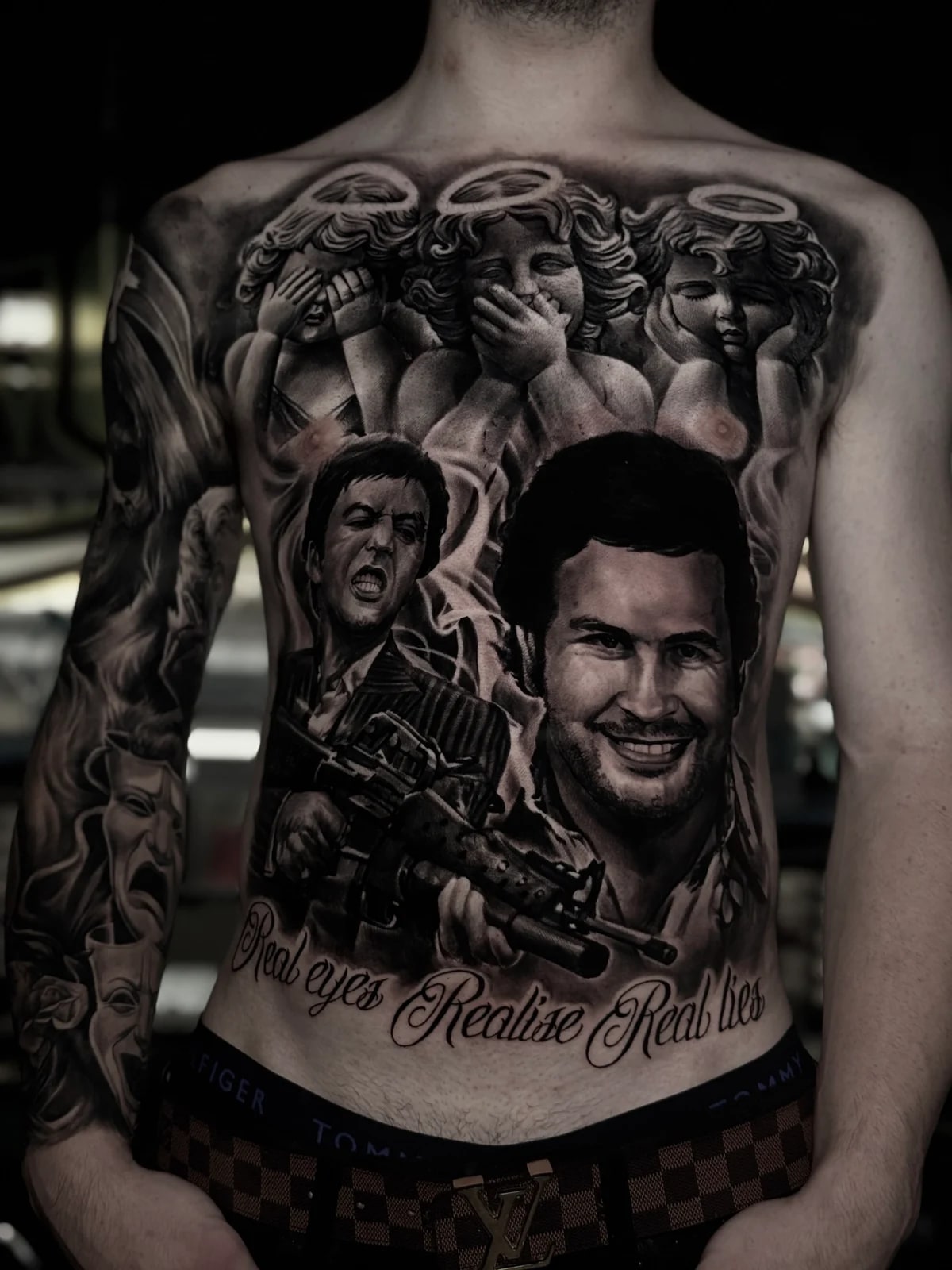 Detailed black and grey realism torso tattoo featuring cherubs, Tony Montana from Scarface, Pablo Escobar, and script reading Real eyes Realise Real lies at Bloodline Tattoo Phuket.