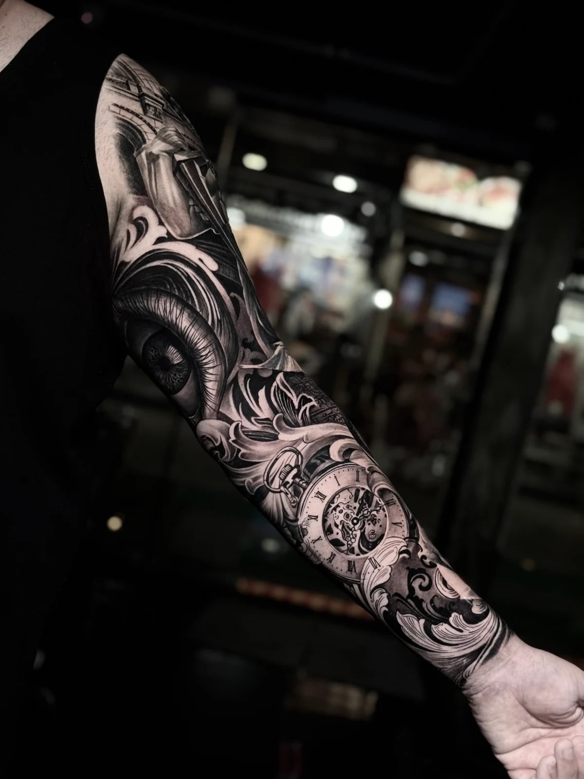 Detailed black and grey realism full sleeve tattoo featuring a realistic human eye, skeleton pocket watch with gears, and ornate filigree scrollwork at Bloodline Tattoo Phuket.