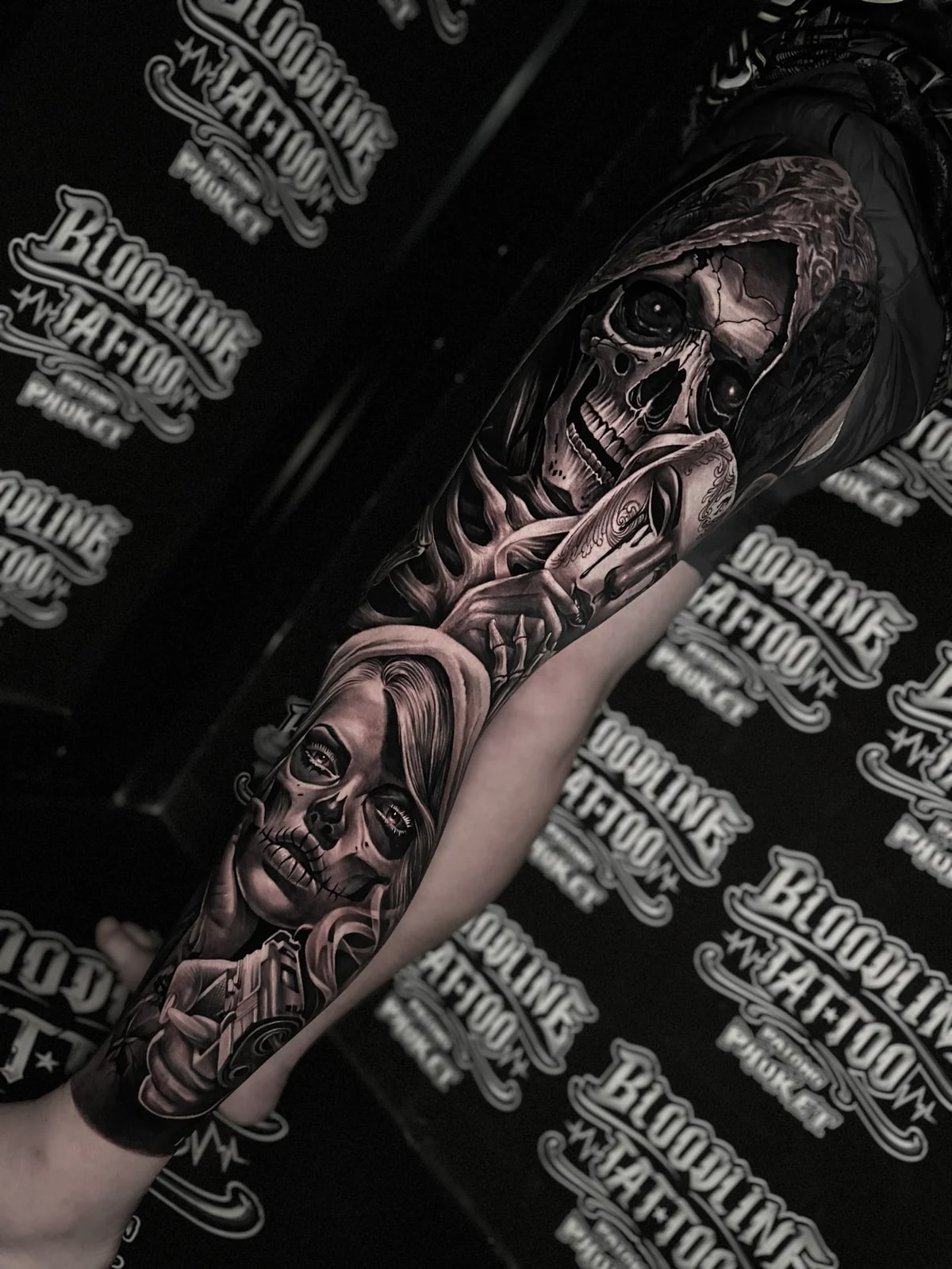 Detailed black and grey realism full leg sleeve tattoo featuring a hooded skull, Venetian mask, Santa Muerte woman, and handgun at Bloodline Tattoo Phuket.
