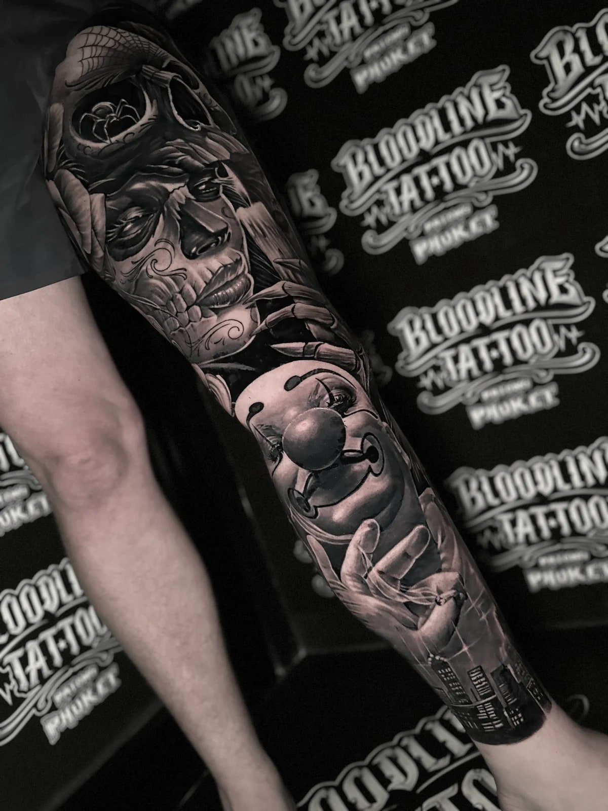 Detailed black and grey realism full leg sleeve tattoo featuring a skull with spider web, sugar skull woman, clown face, smoking hand, and city skyline at Bloodline Tattoo Phuket.