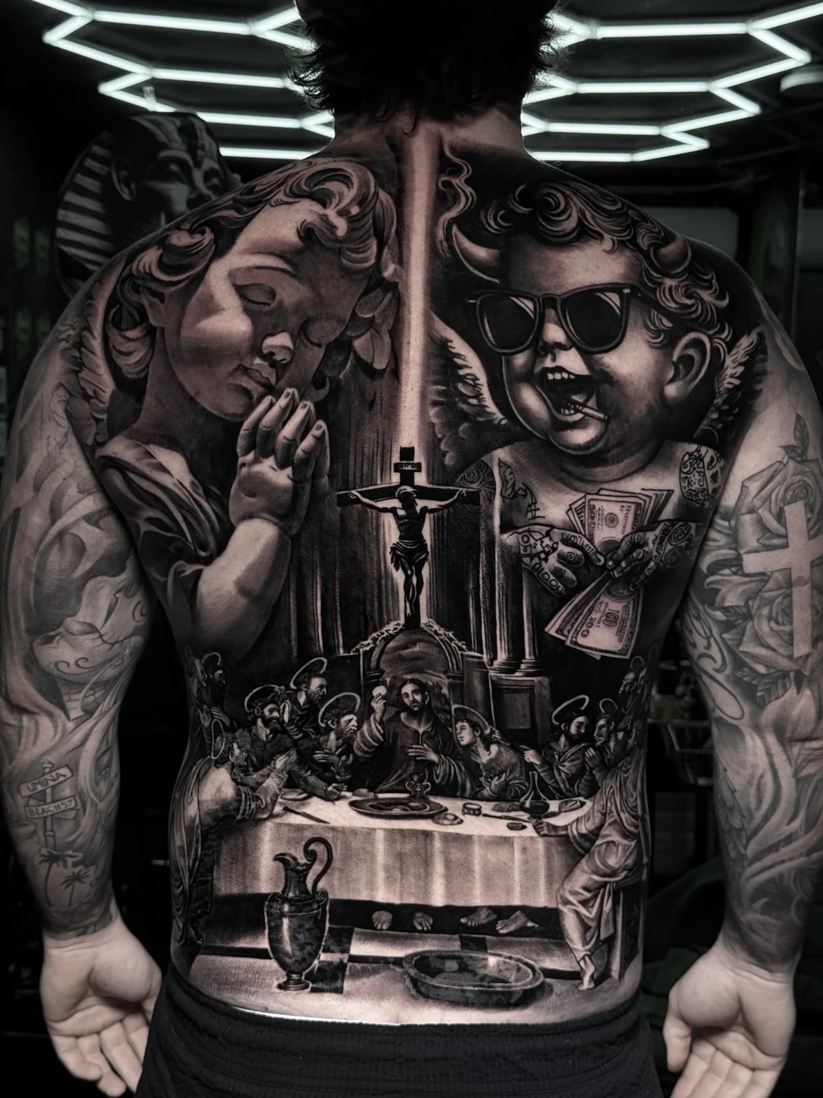 Detailed black and grey realism full back tattoo featuring a praying cherub, devilish cherub with money, crucifix, and The Last Supper at Bloodline Tattoo Phuket.
