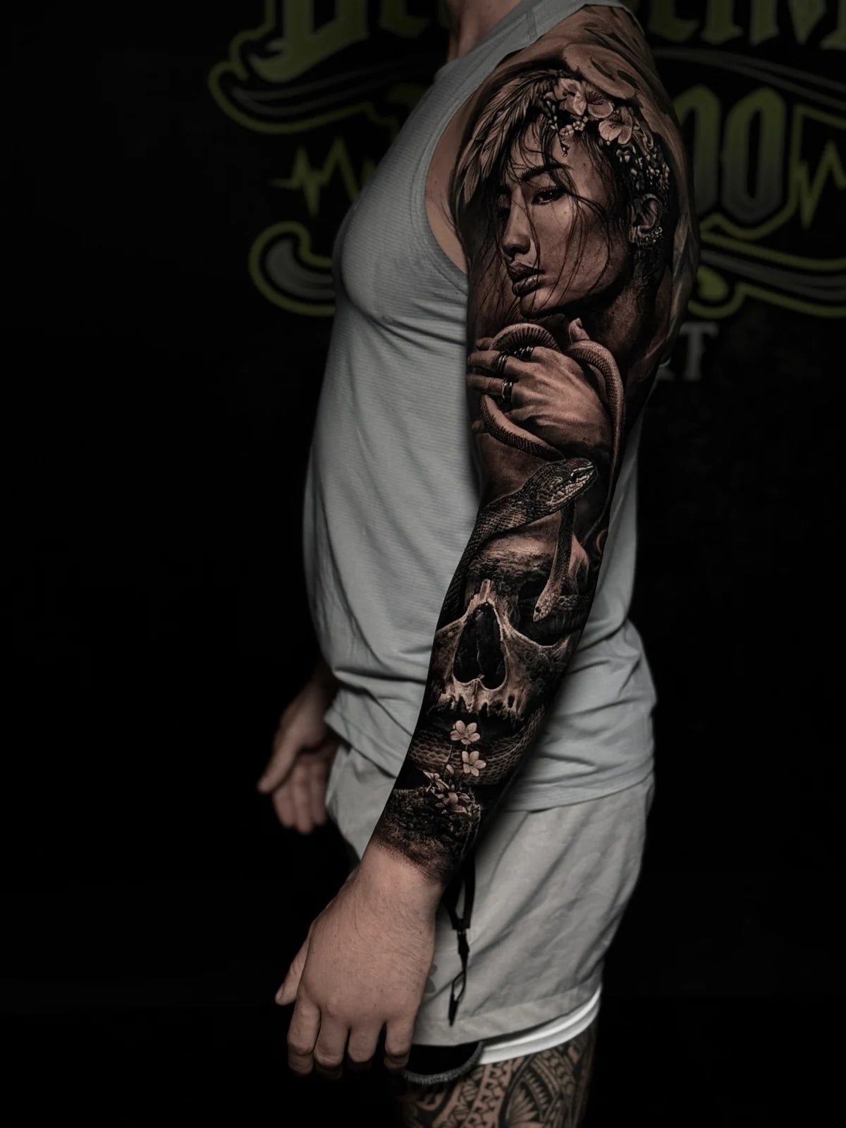 Detailed black and grey realism full sleeve tattoo featuring a woman's floral portrait, hand gripping a snake, realistic skull, and flowers at Bloodline Tattoo Phuket.