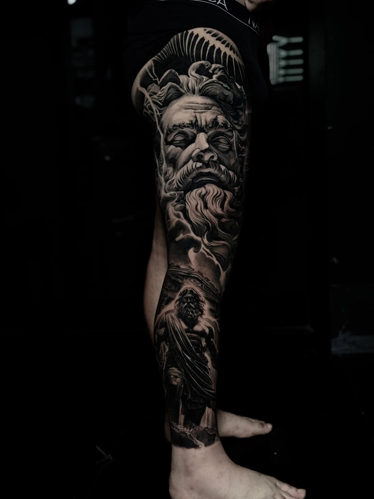 Detailed black and grey realism full leg sleeve tattoo featuring a Greek god portrait on the thigh and a robed figure before classical temple columns at Bloodline Tattoo Phuket.