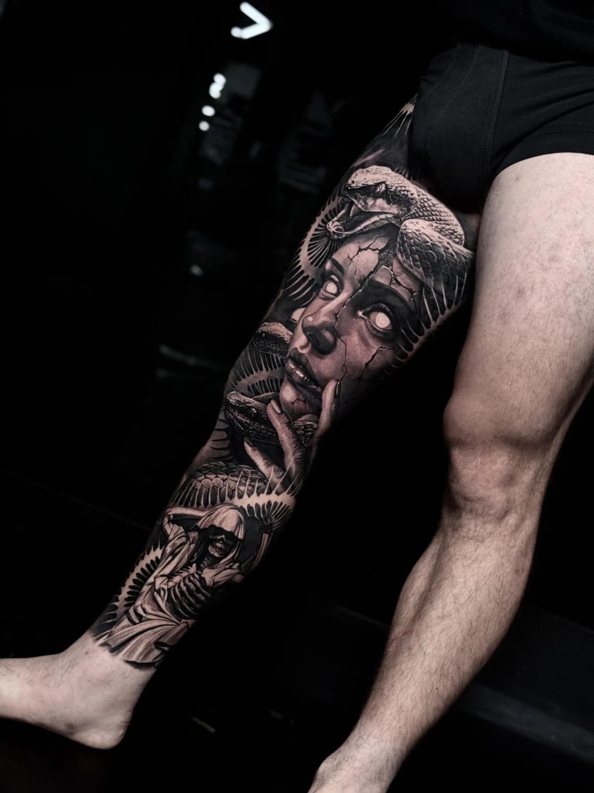 Detailed black and grey realism full leg sleeve tattoo featuring a cracked stone woman with white eyes, coiled snakes, and hooded skeletal reaper at Bloodline Tattoo Phuket.