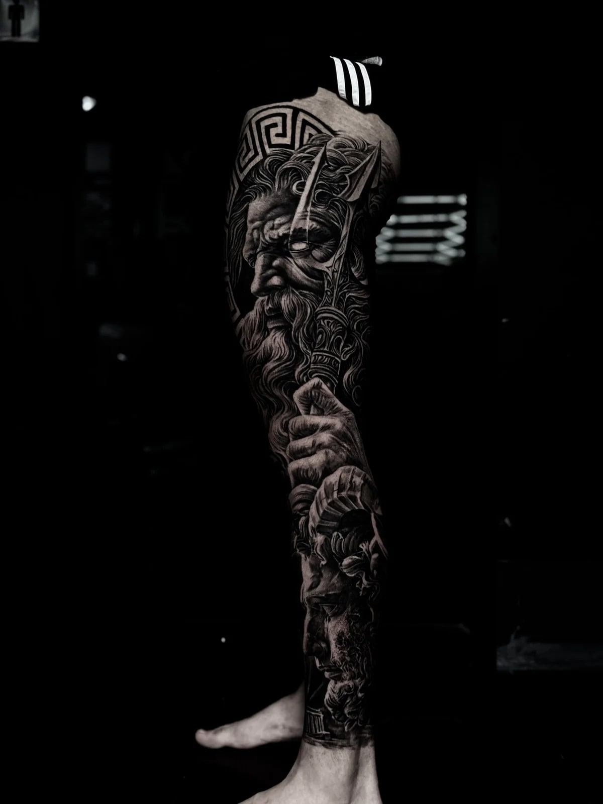 Detailed black and grey realism full leg sleeve tattoo featuring Poseidon with trident, Greek key pattern, and classical temple near the ankle at Bloodline Tattoo Phuket.