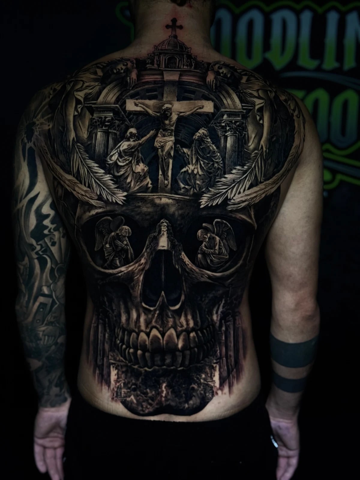 Detailed black and grey realism full back tattoo featuring the Crucifixion in an archway, feathered wings, large skull, and praying angels in the eye sockets at Bloodline Tattoo Phuket.