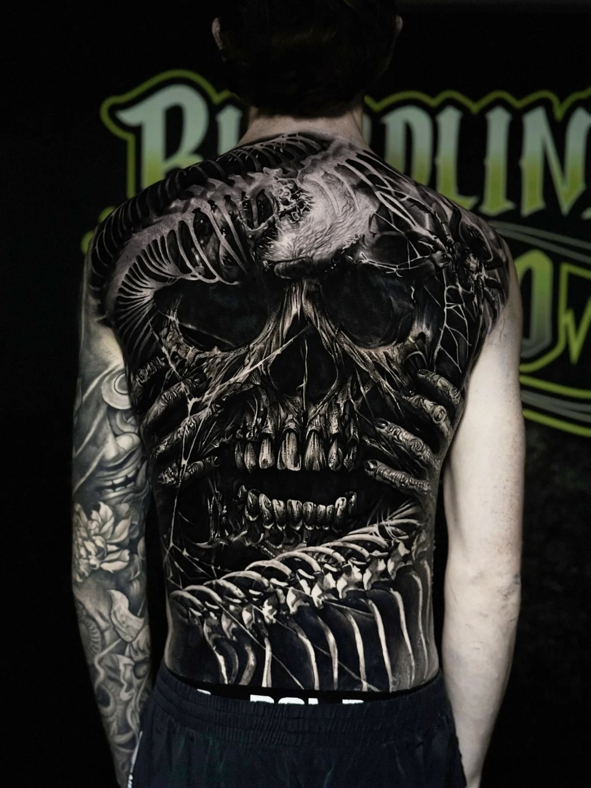 Detailed black and grey realism full back tattoo featuring a massive weathered skull, skeletal gripping hands, ribcage framing, and spider at Bloodline Tattoo Phuket.