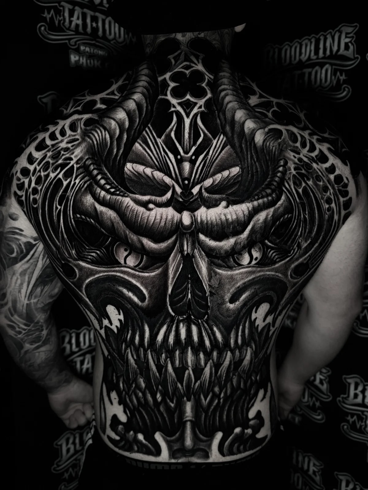 Detailed black and grey realism full back tattoo featuring a horned demonic face with cat-like eyes, jagged teeth, and gothic filigree at Bloodline Tattoo Phuket.