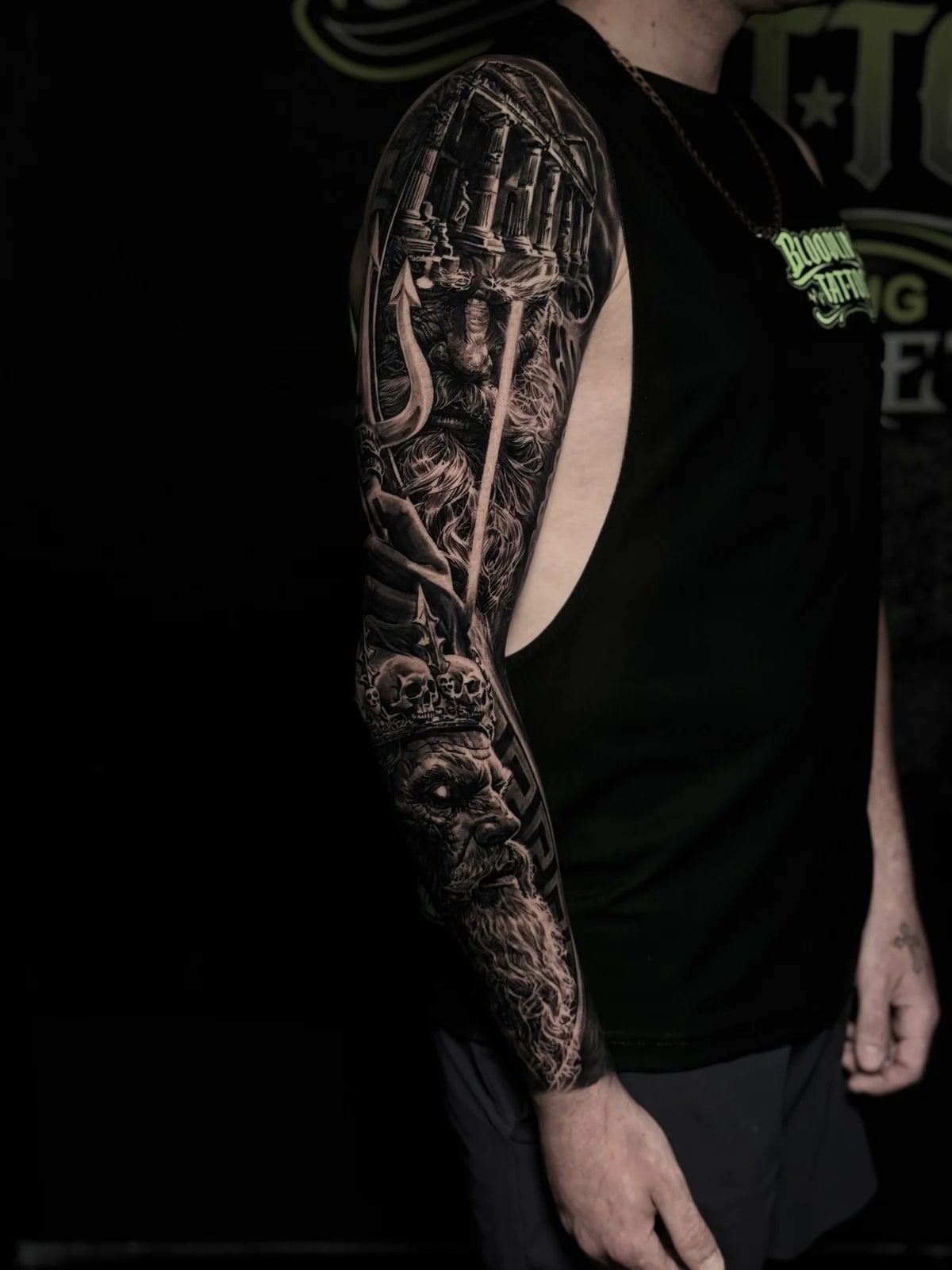 Detailed black and grey realism full sleeve tattoo featuring a Greek temple, Poseidon with trident, and bearded figure with skull crown at Bloodline Tattoo Phuket.