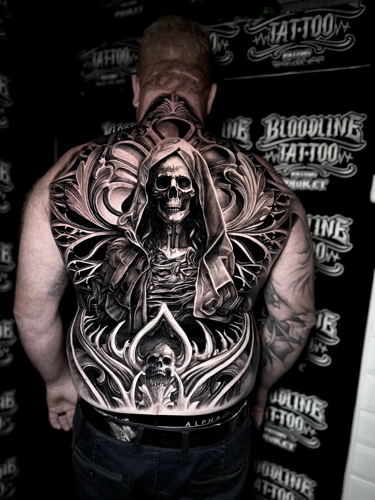 Detailed black and grey realism full back tattoo featuring a hooded skeletal grim reaper, ornate filigree, and screaming skull in a spade frame at Bloodline Tattoo Phuket.